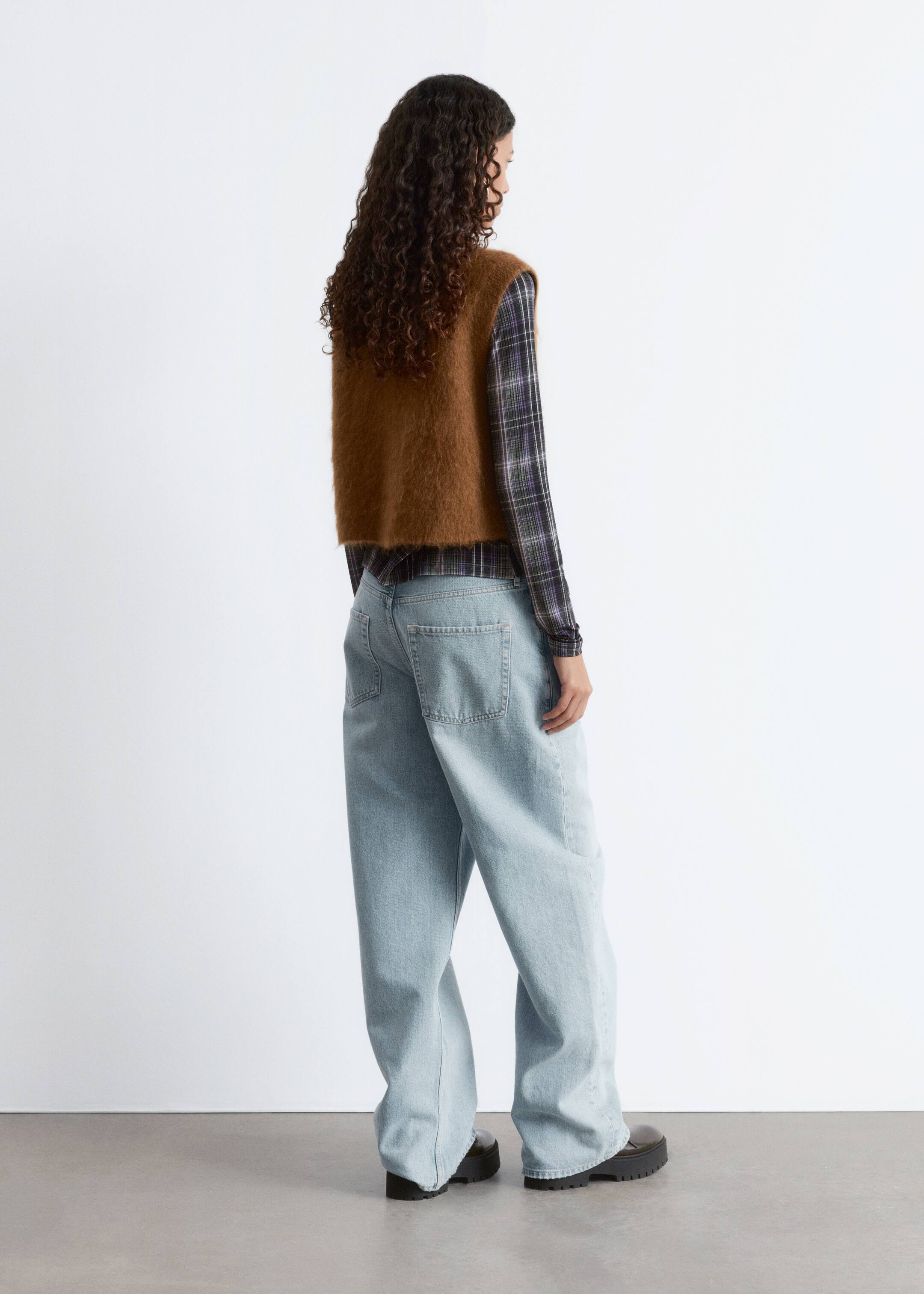 Visa större bild: En outfit presenteras från baksidan och höger sida, med en brun rufsig stickad väst över en mörk rutig långärmad skjorta. Personen har också på sig ljusblå jeans med vida ben och mörka skor med kraftiga sulor.