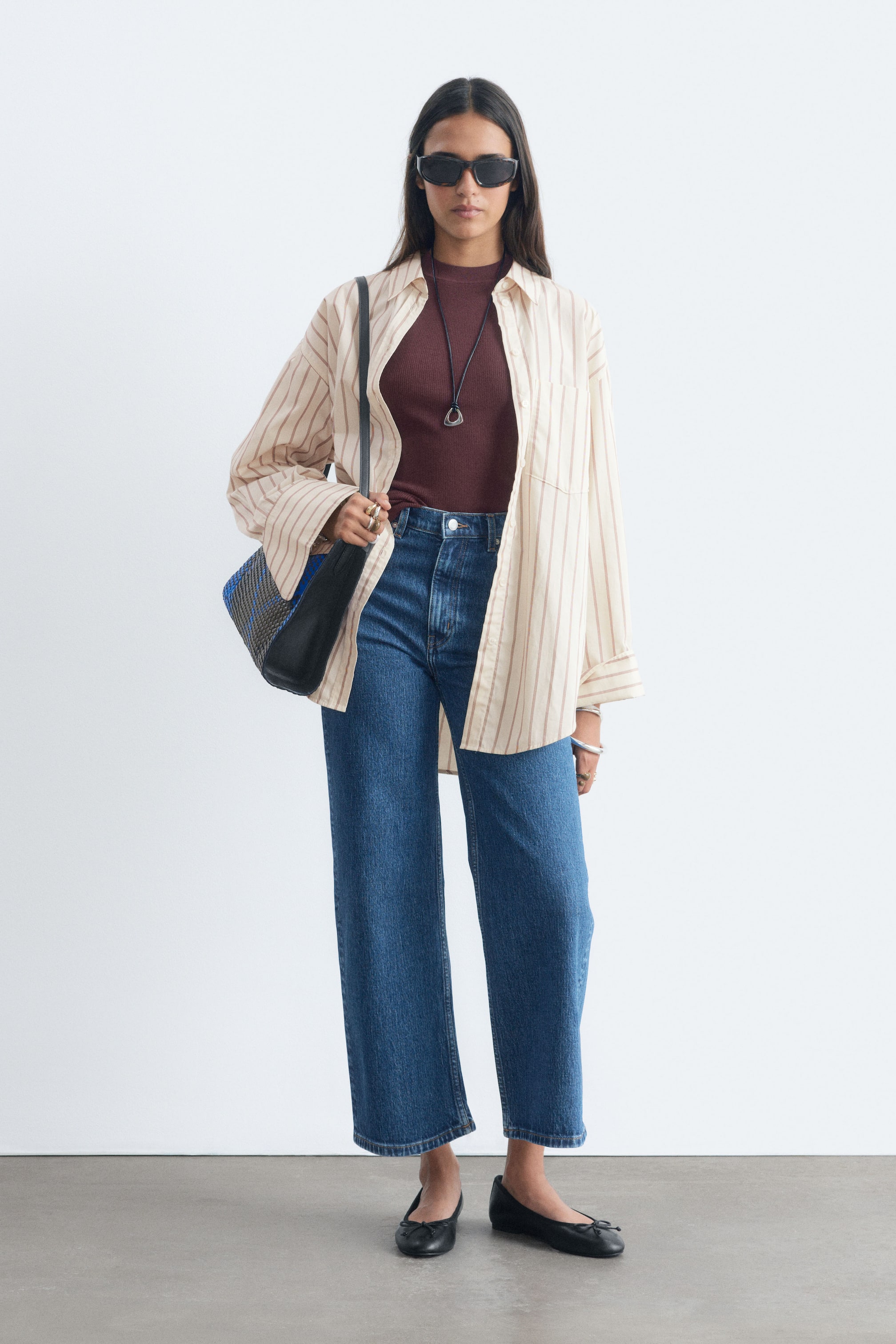 Größeres Bild anzeigen: Der Model trägt hochgeschnittene, dunkelblaue Denim-Jeans mit weitem, verkürztem Bein. Die Jeans verfügen über einen sichtbaren Knopf- und Reißverschlussverschluss und sind mit einem in den Bund gesteckten bordeauxroten Oberteil sowie einem offen getragenen gestreiften Hemd kombiniert.