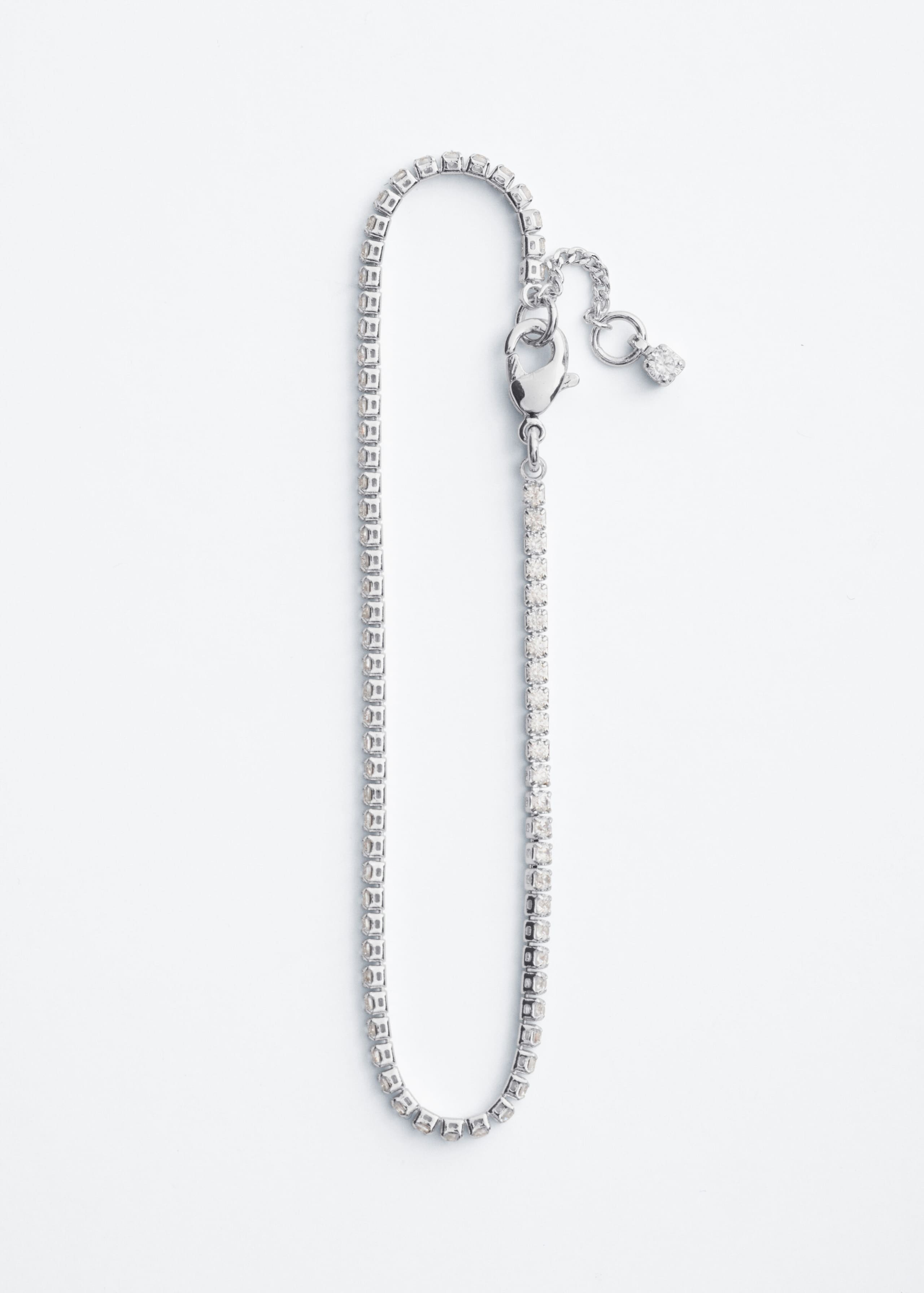 A silver-toned rhinestone tennis bracelet is laid flat in a vertical loop, showcasing a continuous strand of small, closely set rhinestones. The design features a lobster clasp closure and a short extension chain with a single dangling rhinestone charm.