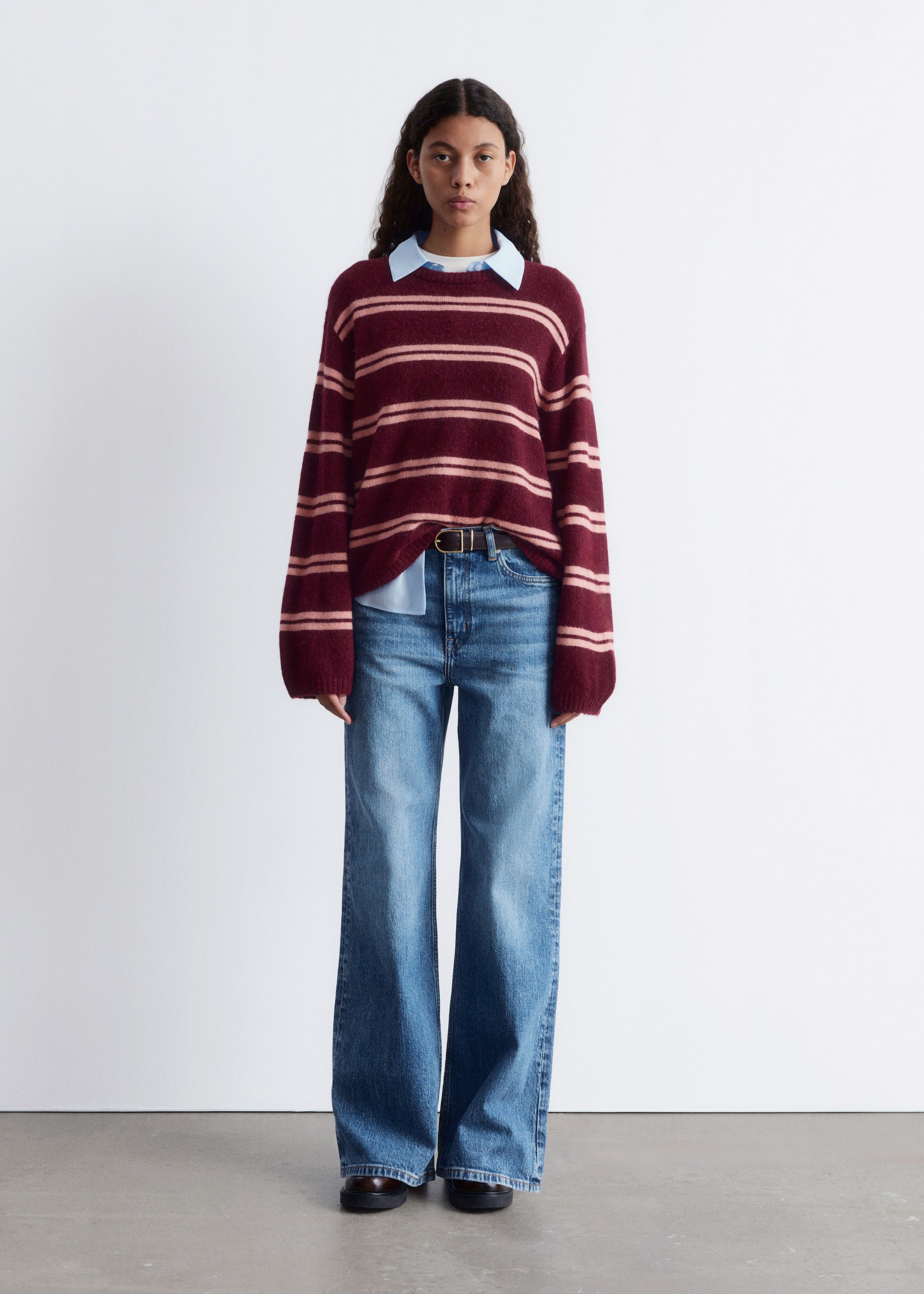 Agrandir l'image: Un pull à col rond rayé bordeaux et rose clair est présenté de face. Le pull est superposé sur une chemise à col bleu clair et est stylé avec un jean bleu à jambes larges, accessoirisé avec une ceinture marron foncé et des mocassins chunky marron foncé.