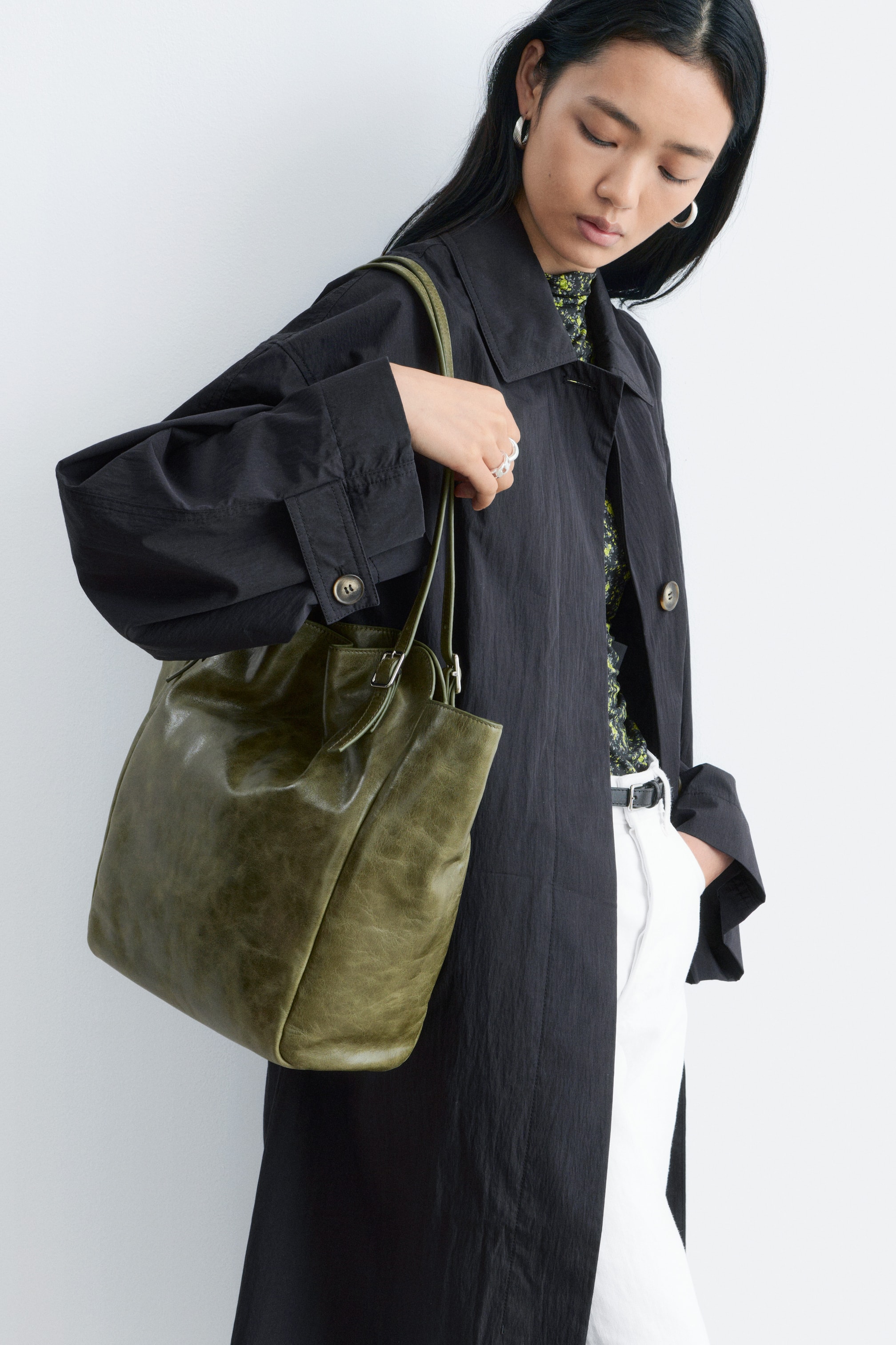 Agrandir l'image: Un sac cabas souple vert olive est tenu par sa seule bandoulière, présentant son profil avant et latéral. Il est doté d'un matériau texturé et froissé avec une ouverture supérieure froncée et une boucle ajustable sur sa bandoulière.