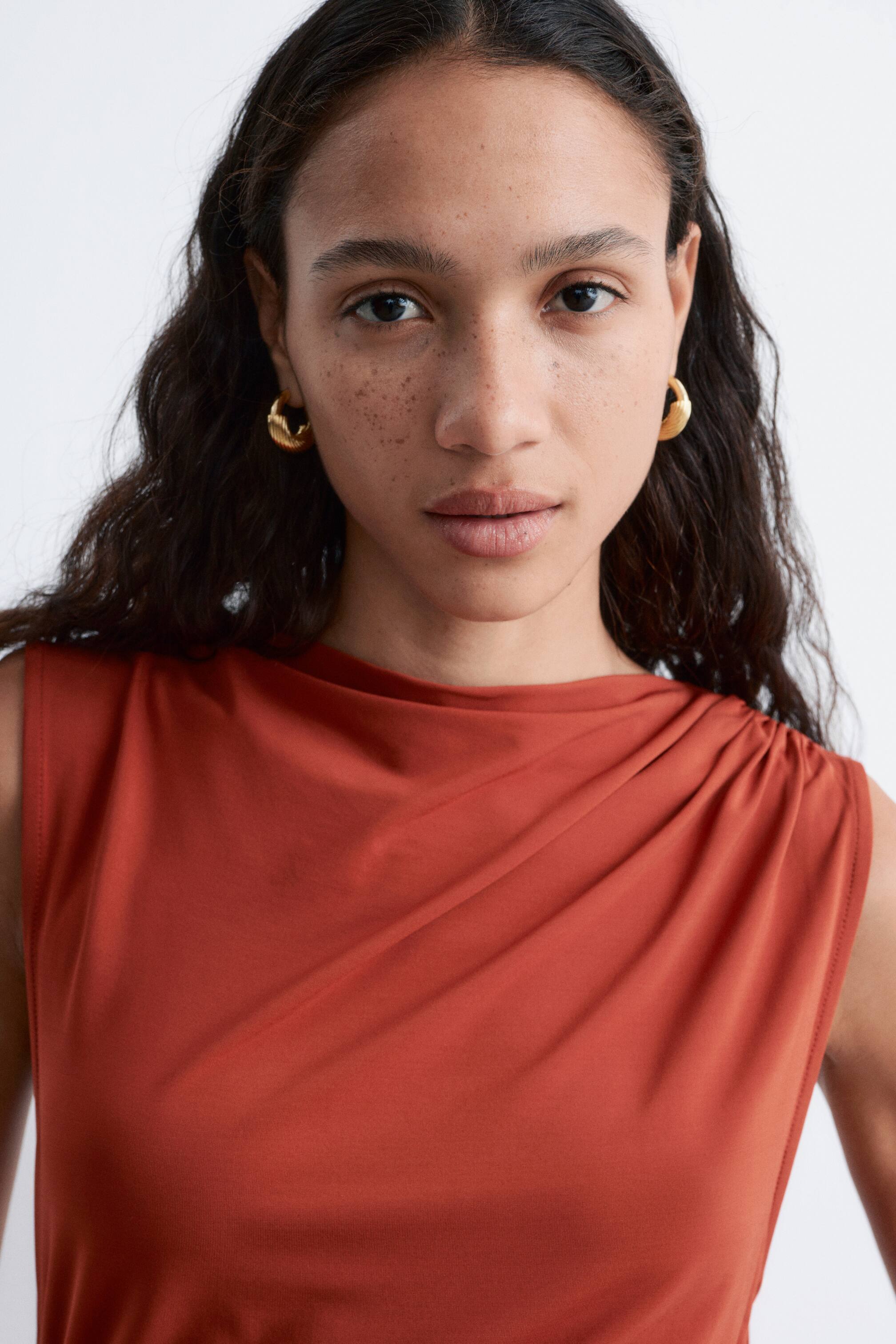 Visualizza immagine più grande: Un top senza maniche color ruggine presenta uno scollo drappeggiato. Il tessuto si raccoglie sulla spalla destra di chi lo indossa, creando morbide pieghe sulla parte anteriore.