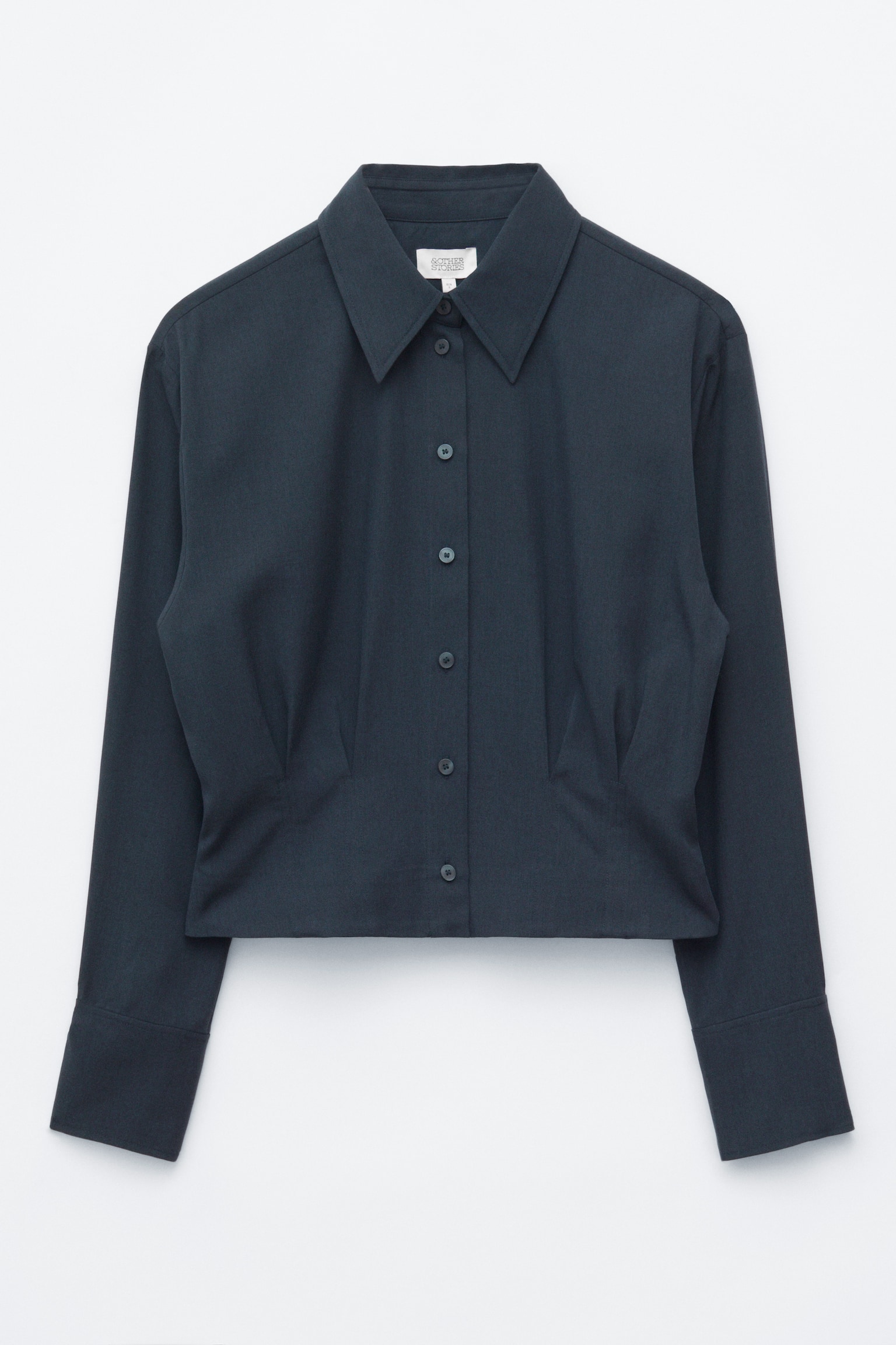 Uma camisa cinzento-escura de manga comprida está virada para a frente, destacando uma gola pontiaguda definida, uma frente com botões e mangas compridas com punhos largos. A camisa apresenta pregas estruturadas na cintura que definem a sua silhueta e uma bainha curta.
