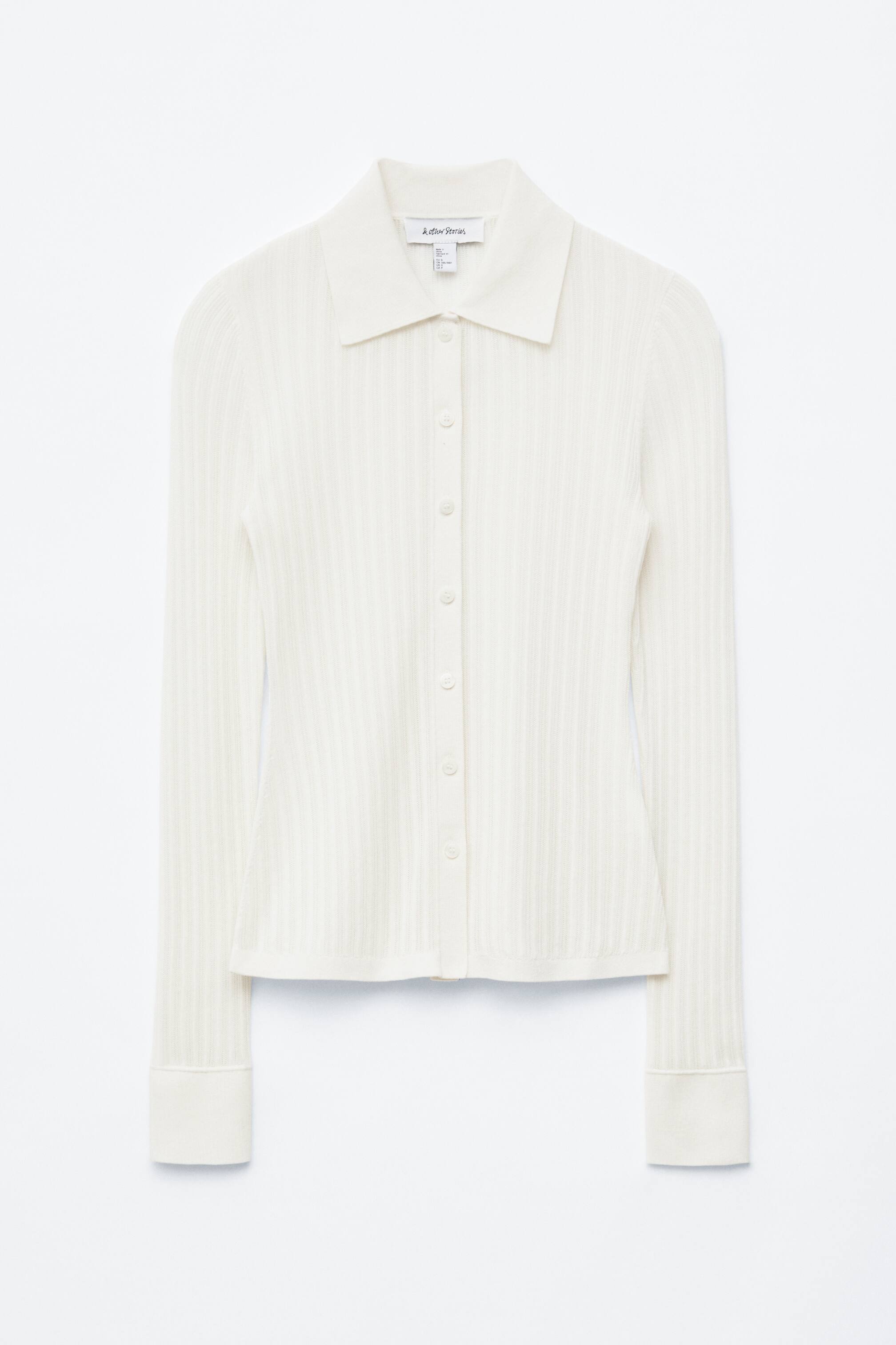Zobrazit větší obrázek: Krémově zbarvený žebrovaný pletený top je prezentován zepředu, s výrazným špičatým límcem a plnou knoflíkovou légou s kulatými knoflíky uprostřed. Dlouhé rukávy končí širokými, přeloženými manžetami.