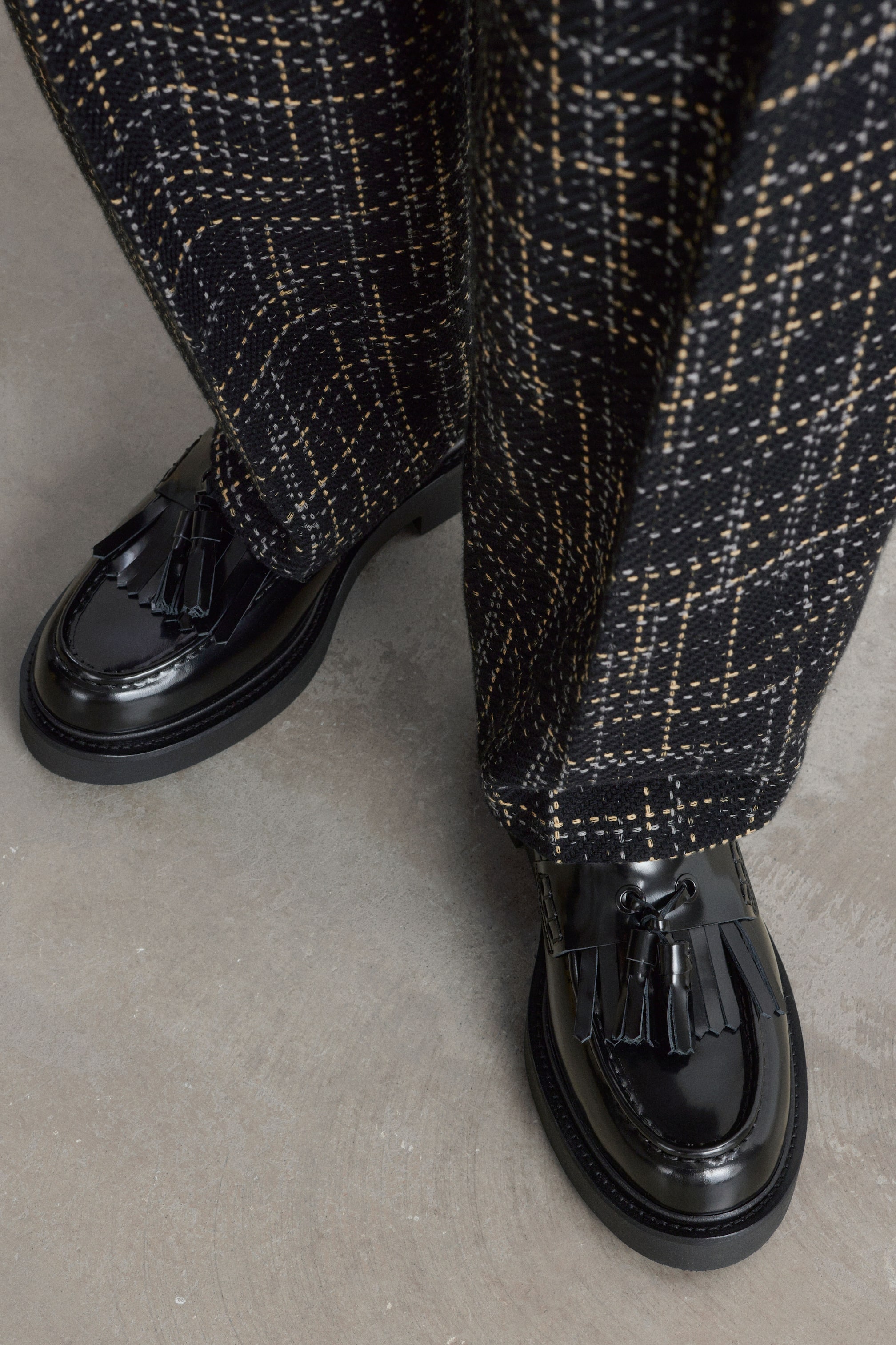 Größeres Bild anzeigen: Der Model trägt schwarze, polierte Leder-Loafer mit Kiltie-Fransen und Quasten. Die Schuhe mit grober Sohle sind mit dunklen, strukturierten Karohosen kombiniert.