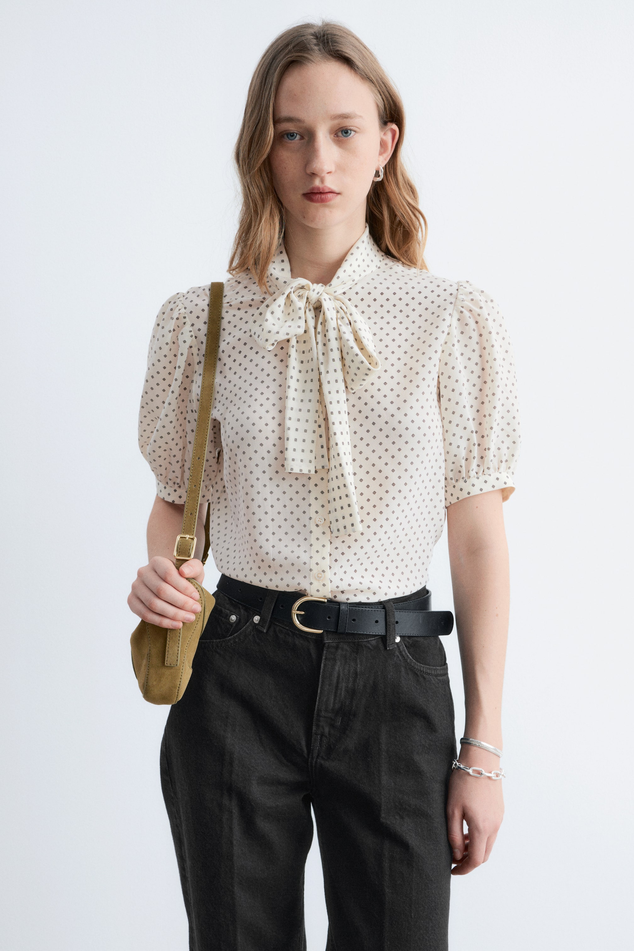 Agrandir l'image: La blouse blanc cassé présente de petits motifs carrés foncés et un col lavallière noué en un nœud soigné. Elle est conçue avec des manches courtes et bouffantes et se ferme par des boutons sur le devant.