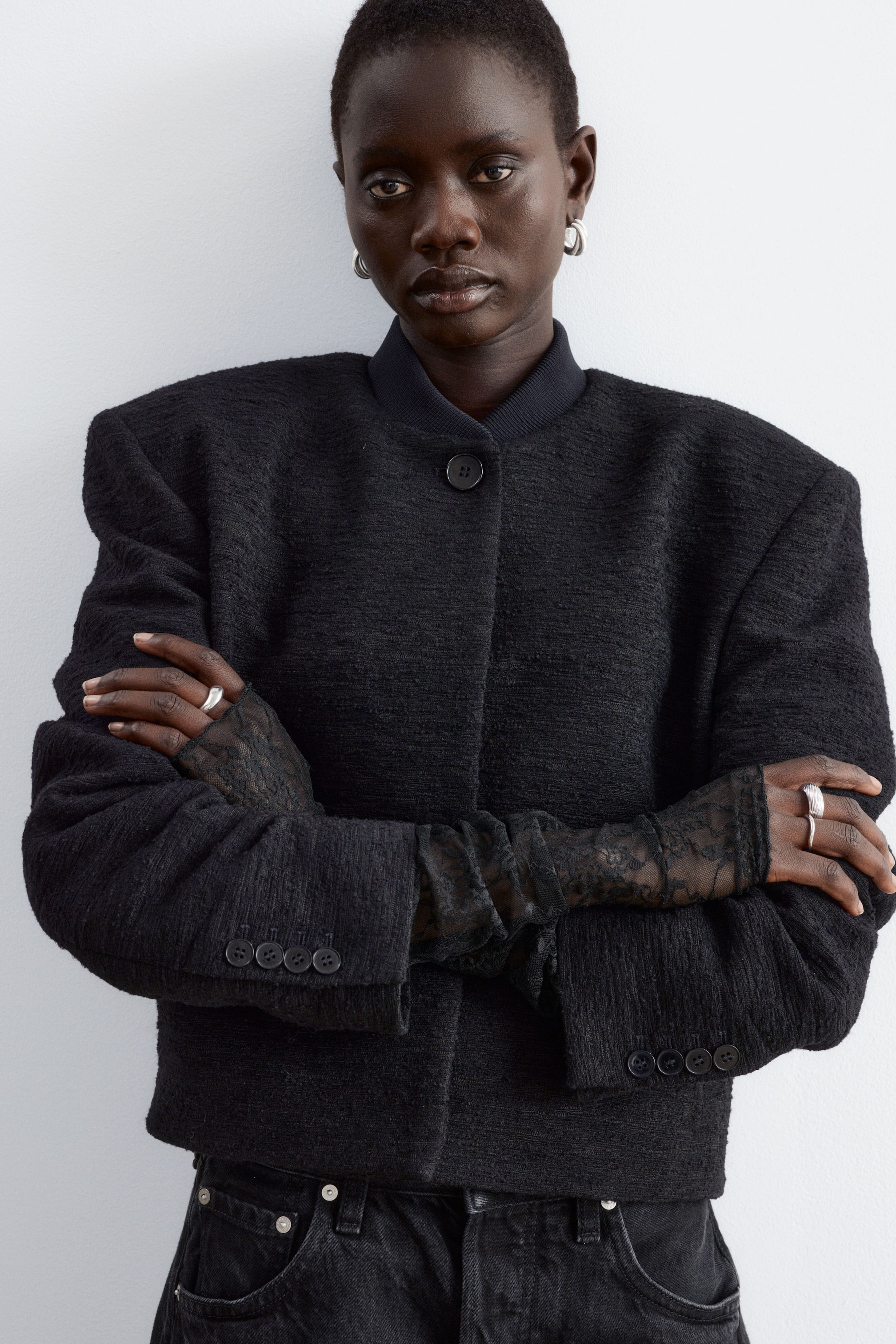 View larger image: Black lace fingerless gloves are being worn by the model, extending along the arms and layered beneath the sleeves of a textured black jacket. The elongated gloves cover the hands while leaving the fingers exposed as the model stands with crossed arms.