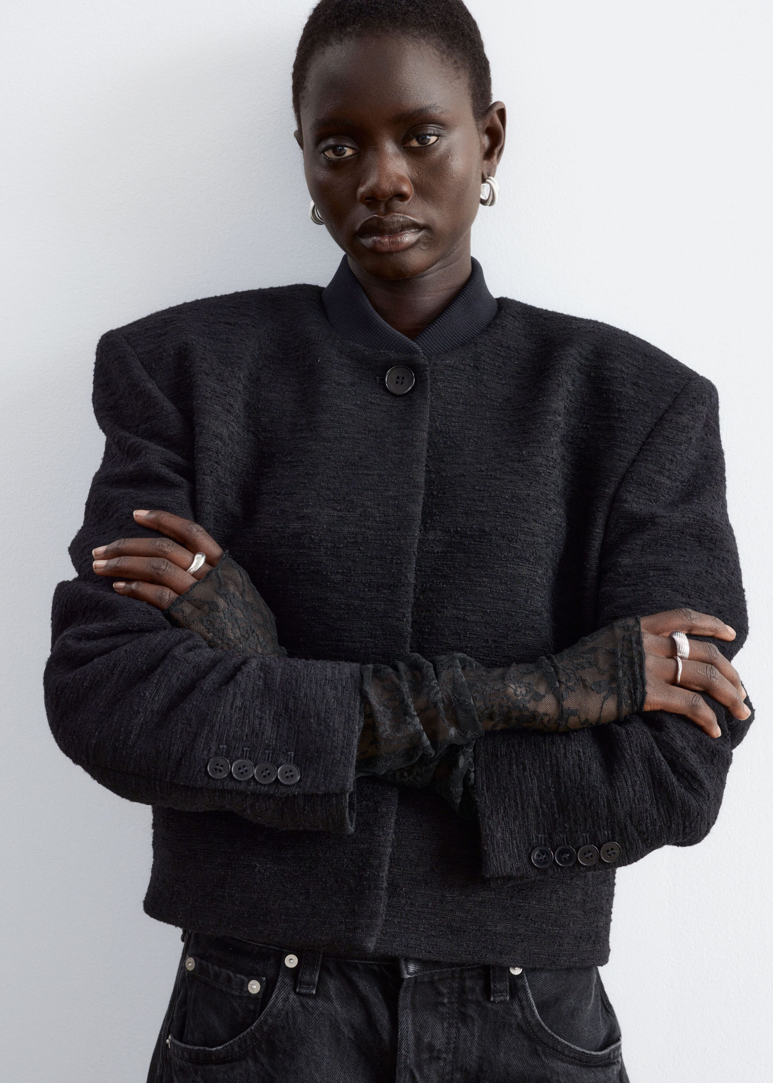 Black lace fingerless gloves are being worn by the model, extending along the arms and layered beneath the sleeves of a textured black jacket. The elongated gloves cover the hands while leaving the fingers exposed as the model stands with crossed arms.