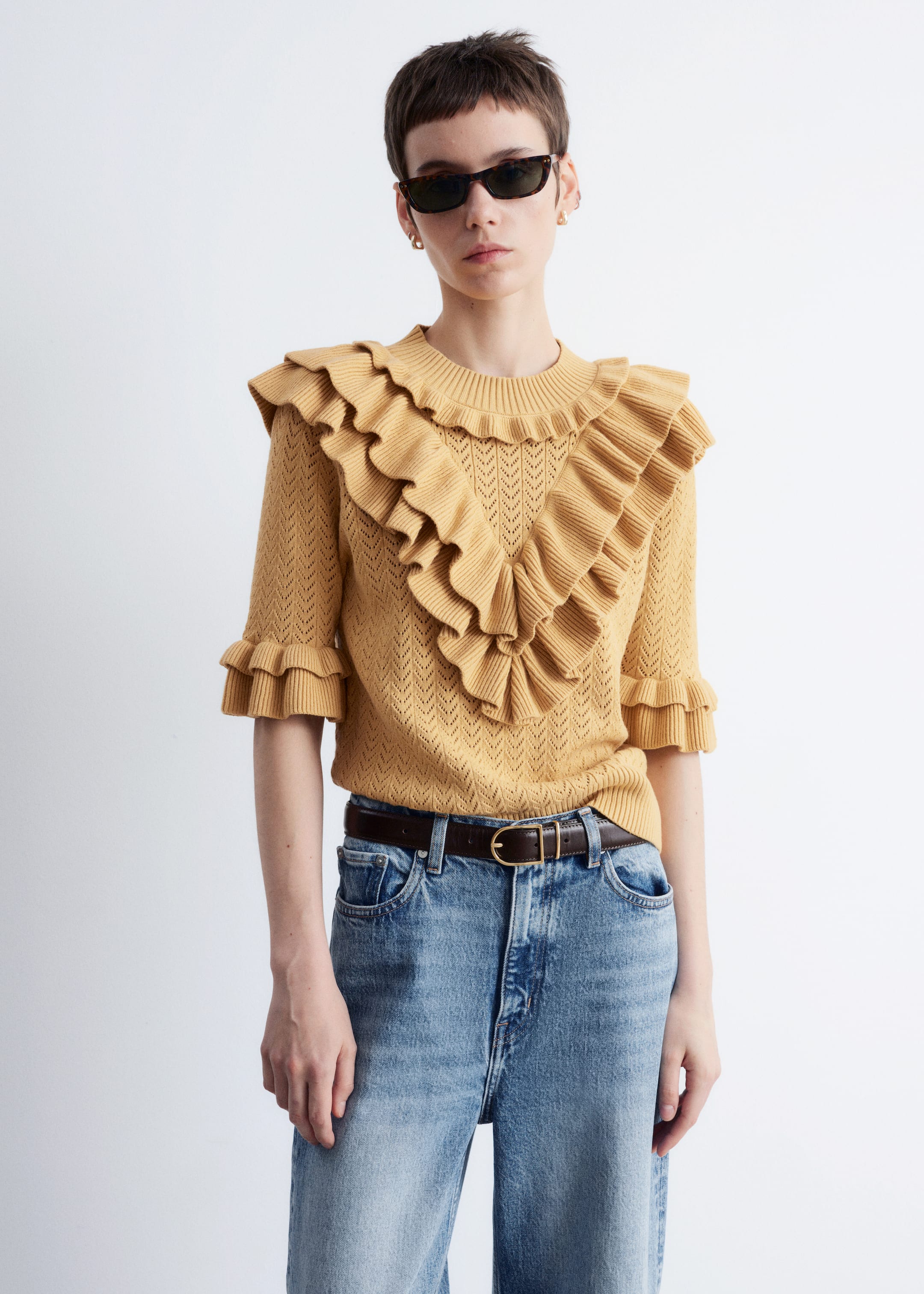 Ver imagem maior: Um top de malha pointelle em amarelo mostarda está a ser usado pela modelo. Apresenta uma gola redonda canelada, mangas curtas com punhos franzidos e folhos em camadas que contornam a gola e formam um V na frente. O top é combinado com jeans de lavagem clara e um cinto castanho.