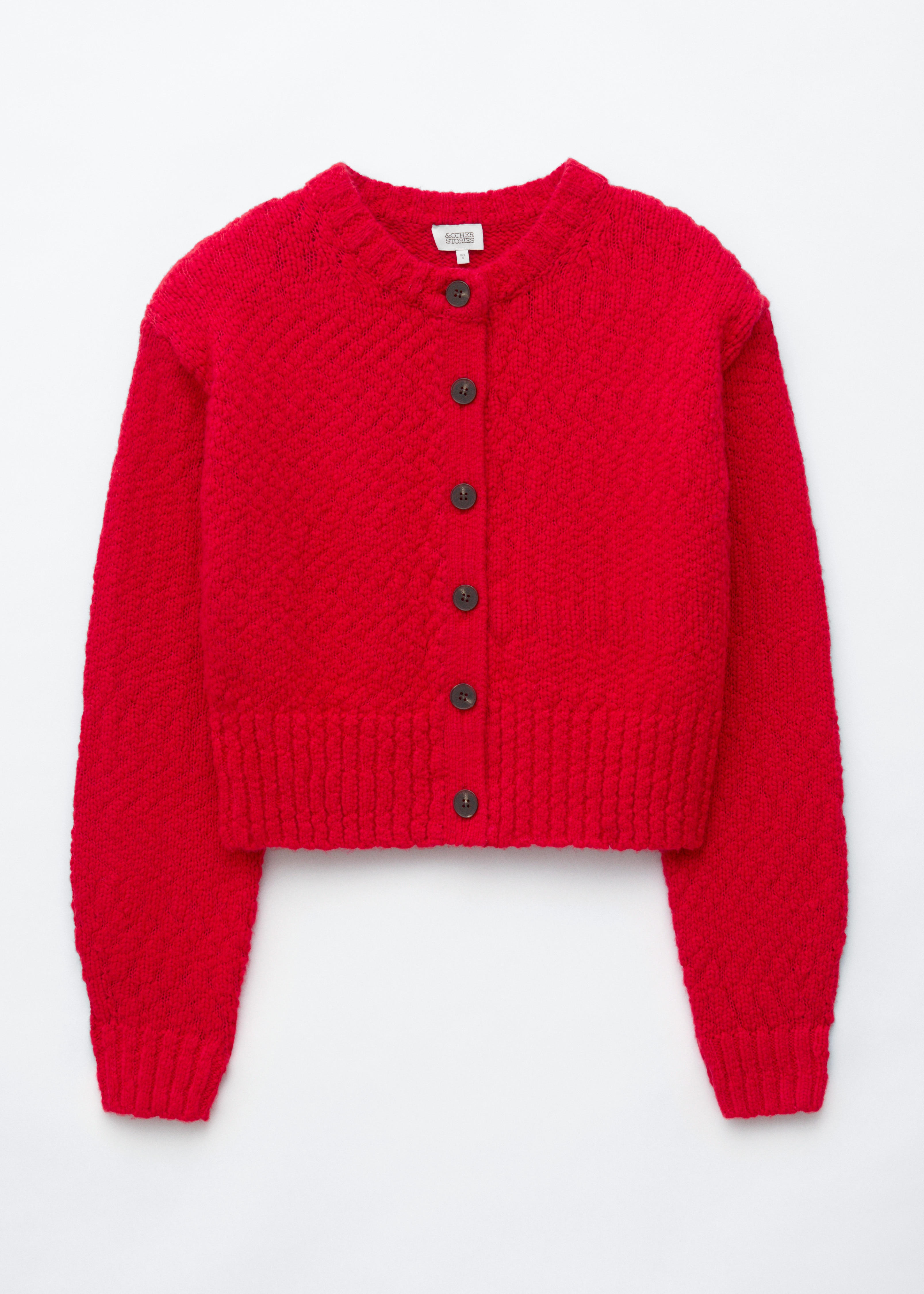 A red wool-cotton cardigan featuring a textured knit, front button fastening with dark buttons, and a ribbed crew neck is worn by the model. The cardigan is styled with blue jeans, with the modelâs hands resting in the front trouser pockets.