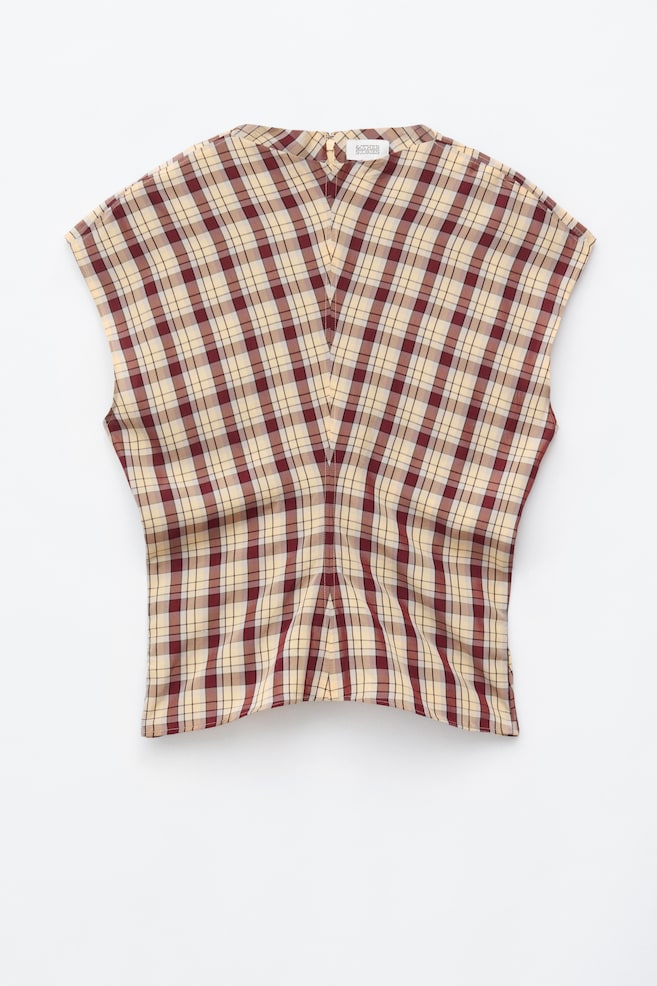 La blusa de cuadros muestra un patrón de cuadros en crema, burdeos y negro. Presenta mangas tipo capa y un escote alto, ligeramente redondeado, con una costura central en forma de V que recorre el frente y crea una silueta entallada. - 1