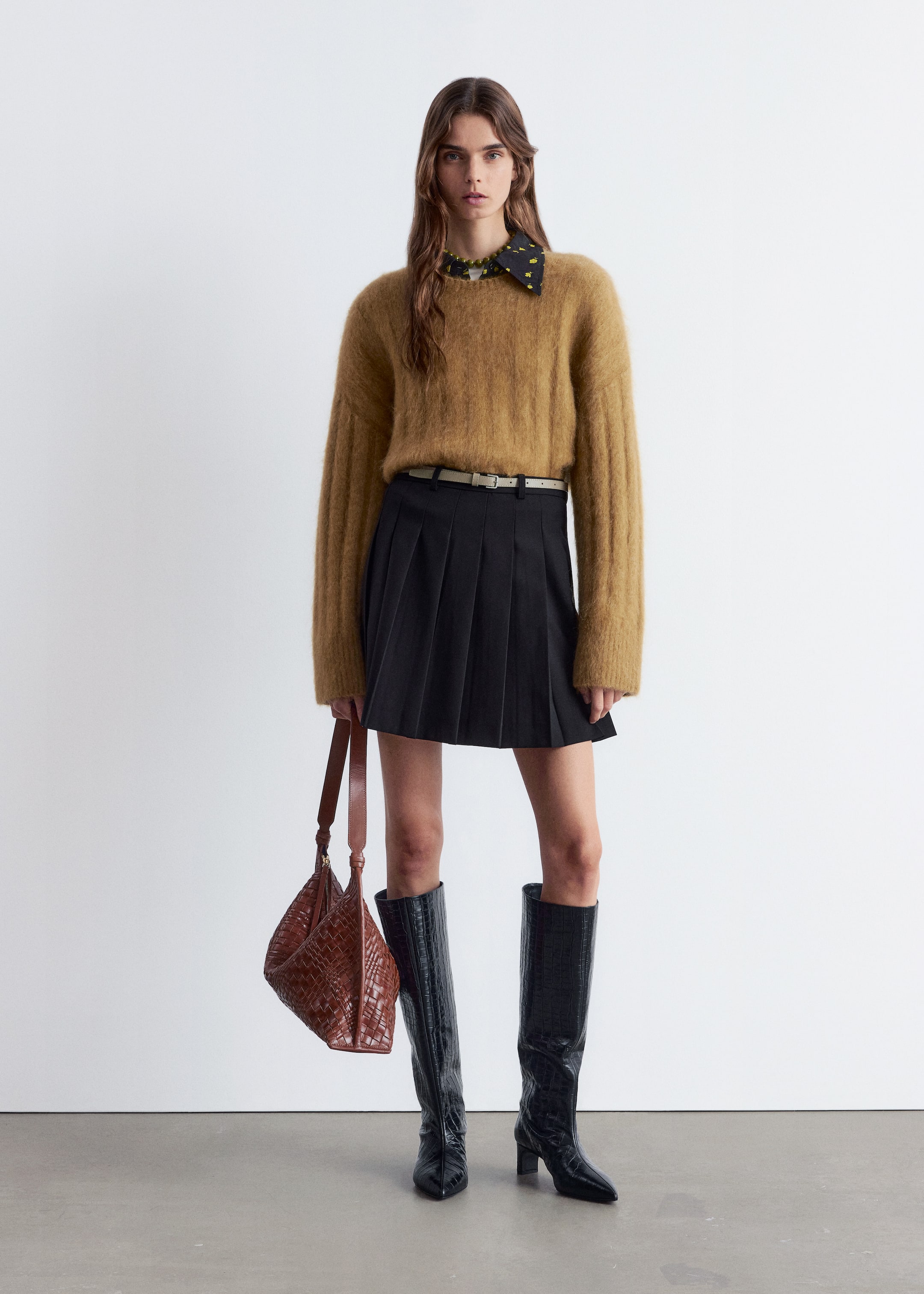 Agrandir l'image: Une tenue est présentée, avec un pull à col rond en tricot duveteux de couleur camel avec des manches longues, superposé sur une chemise noire à col avec un motif floral jaune et un bord à volants visible. Une mini-jupe plissée noire est portée avec une fine ceinture de couleur claire. Pour compléter l'ensemble, des bottes noires hauteur genou avec une finition texturée et un sac hobo tressé marron est tenu par sa sangle.