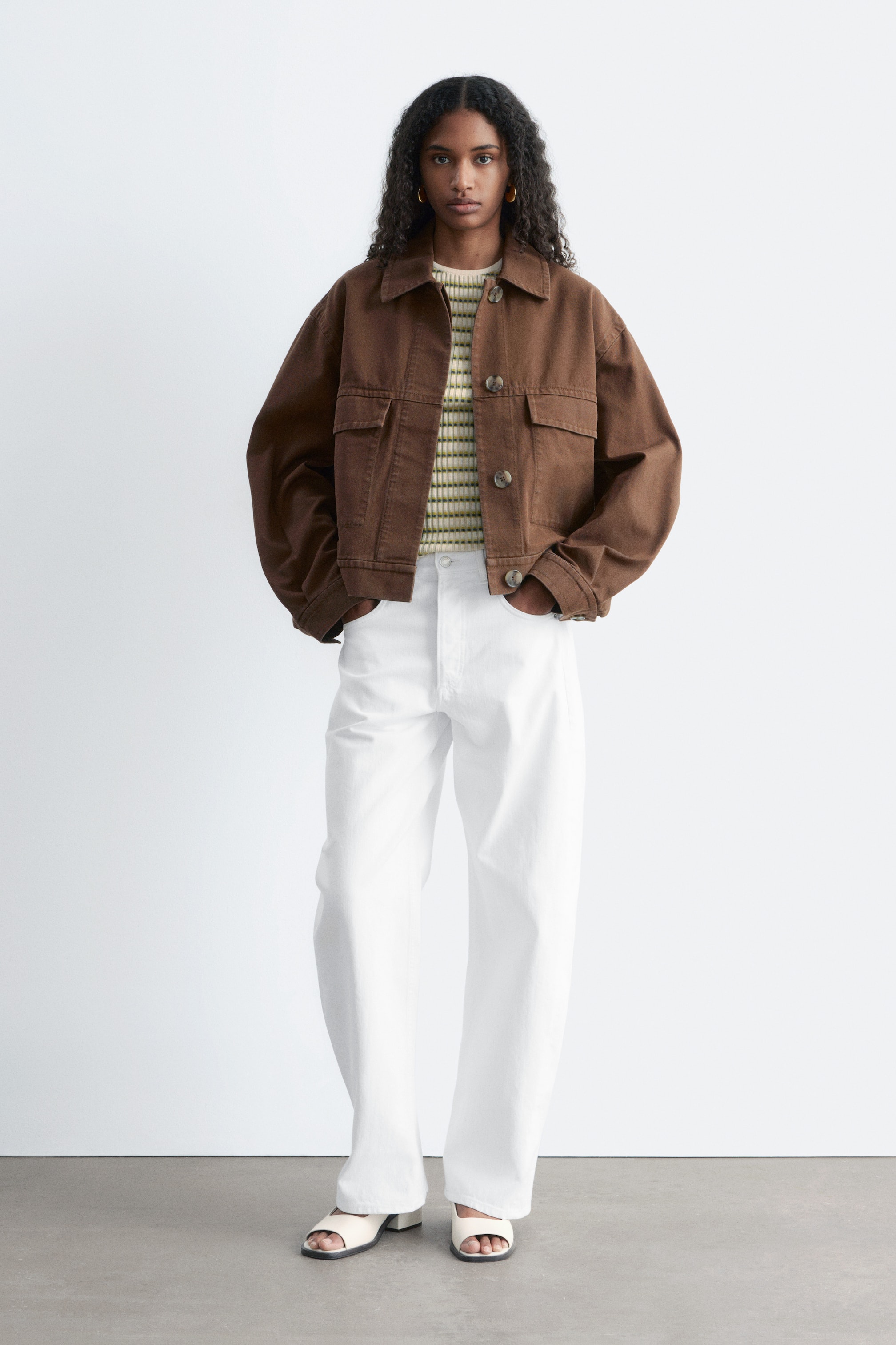 Größeres Bild ansehen: Ein brauner, schwerer Baumwolljacke wird vom Model getragen, offen über einem gestreiften Oberteil. Sie zeichnet sich durch eine kastige Silhouette, zwei Brusttaschen mit Klappen, sichtbare Knöpfe an der Vorderseite und markante Schnittlinien aus.