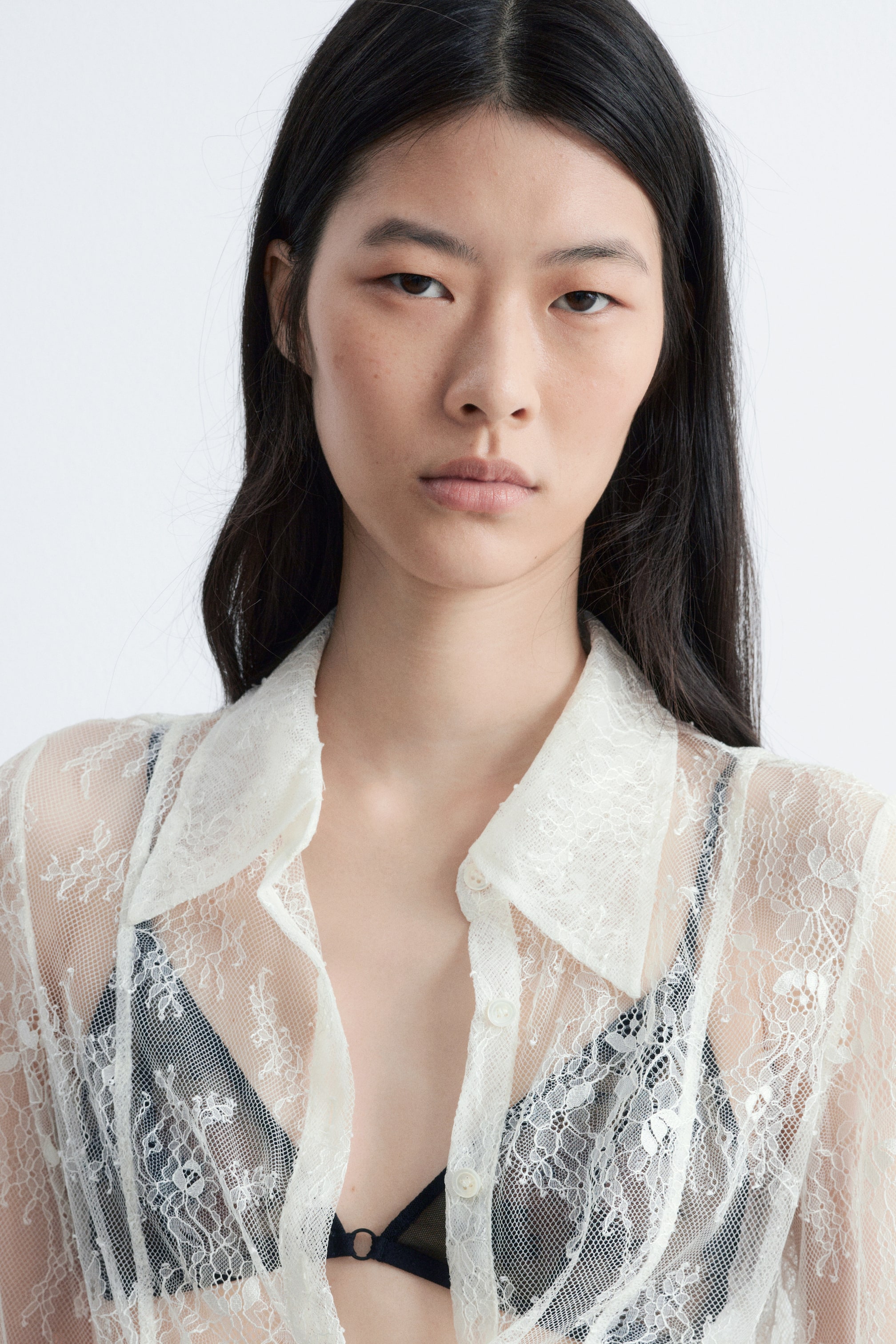 Visualizza immagine più grande: Una camicia trasparente in pizzo color avorio con bottoni è indossata, caratterizzata da un colletto a punta e un delicato motivo floreale. La camicia è parzialmente sbottonata sul davanti, rivelando un reggiseno a fascia nero sotto.