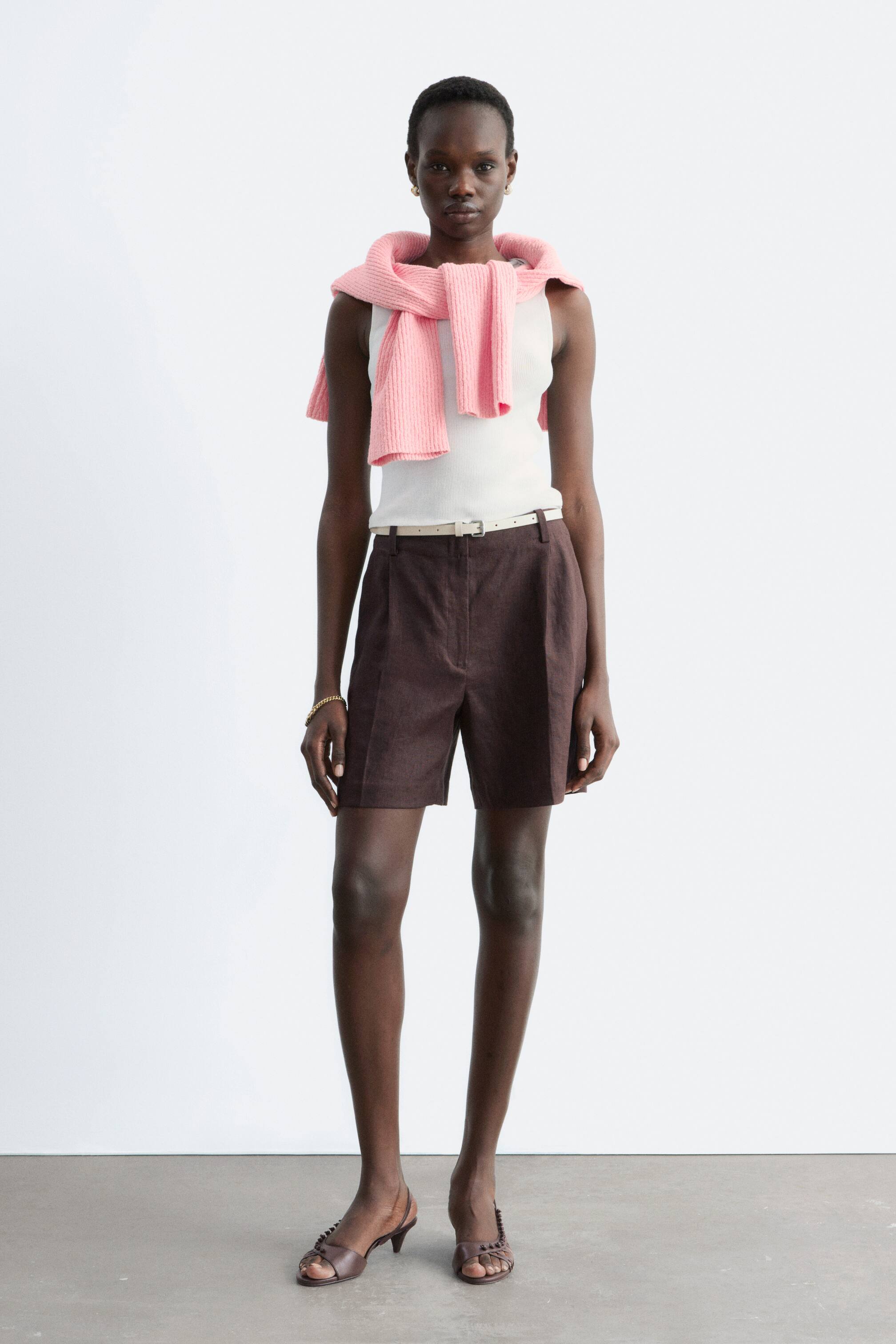 Ver imagem maior: Calções plissados em castanho escuro apresentam cintura alta com presilhas para cinto, fecho frontal com botão e fecho éclair, e bolsos laterais. Atingem acima do joelho e são usados com um cinto bege claro.