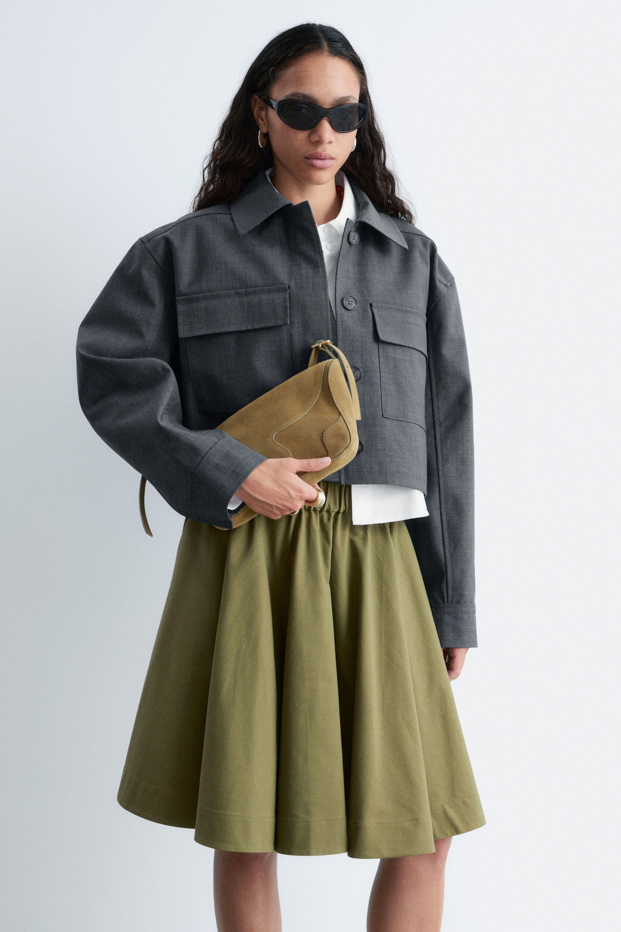 Προβολή μεγαλύτερης εικόνας: Ένα κοντό μπουφάν σε σκούρο γκρι χρώμα είναι στραμμένο προς τα εμπρός, αναδεικνύοντας έναν μυτερό γιακά, δύο τσέπες στο στήθος με καπάκι και κουμπιά, και μακριά μανίκια. Παρουσιάζει μια χαλαρή, κοντή εφαρμογή και είναι φορεμένο πάνω από ένα λευκό πουκάμισο.