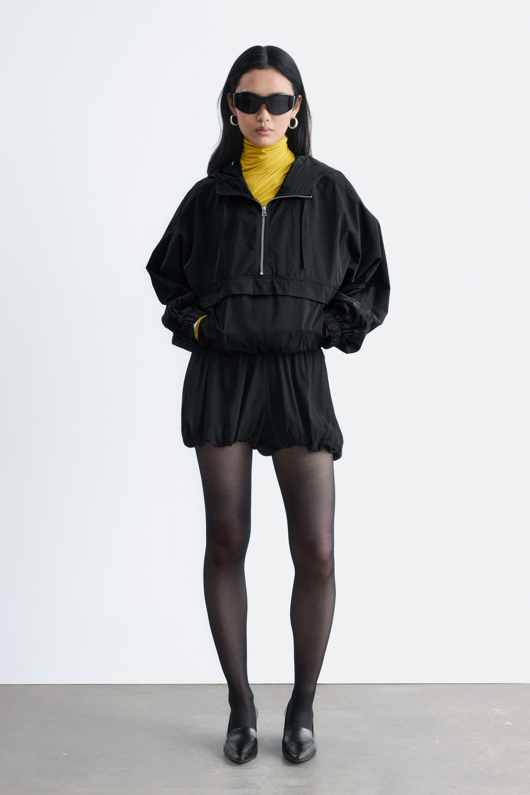 Agrandir l'image: Une veste anorak noire est dotée d'une capuche, d'une demi-fermeture éclair à l'avant et de poignets et ourlet élastiqués, créant une silhouette volumineuse, et est superposée sur un col roulé jaune. Elle est associée à un short noir assorti, qui présente une taille élastiquée et des jambes amples et volumineuses.