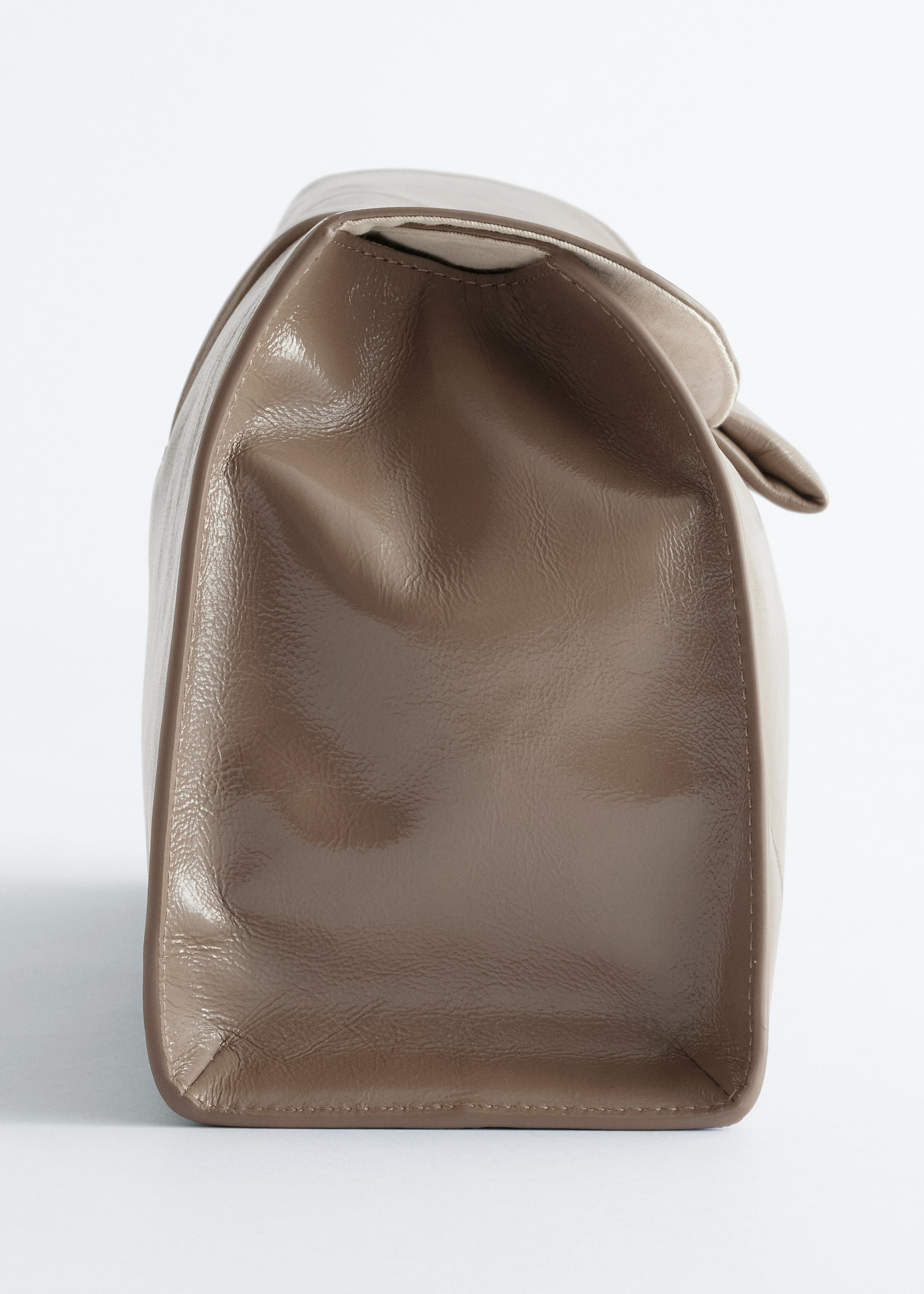 Größeres Bild ansehen: Eine weiche, taupefarbene Ledertasche wird von der Seite gezeigt, wodurch ihr geschmeidiges Material, der flache Boden und die sichtbaren Nähte, die die Struktur der Tasche hervorheben, zu sehen sind. Das Leder zeigt ein halbglänzendes Finish mit natürlichen Falten, und ein Teil der oberen Klappe biegt sich nach unten.