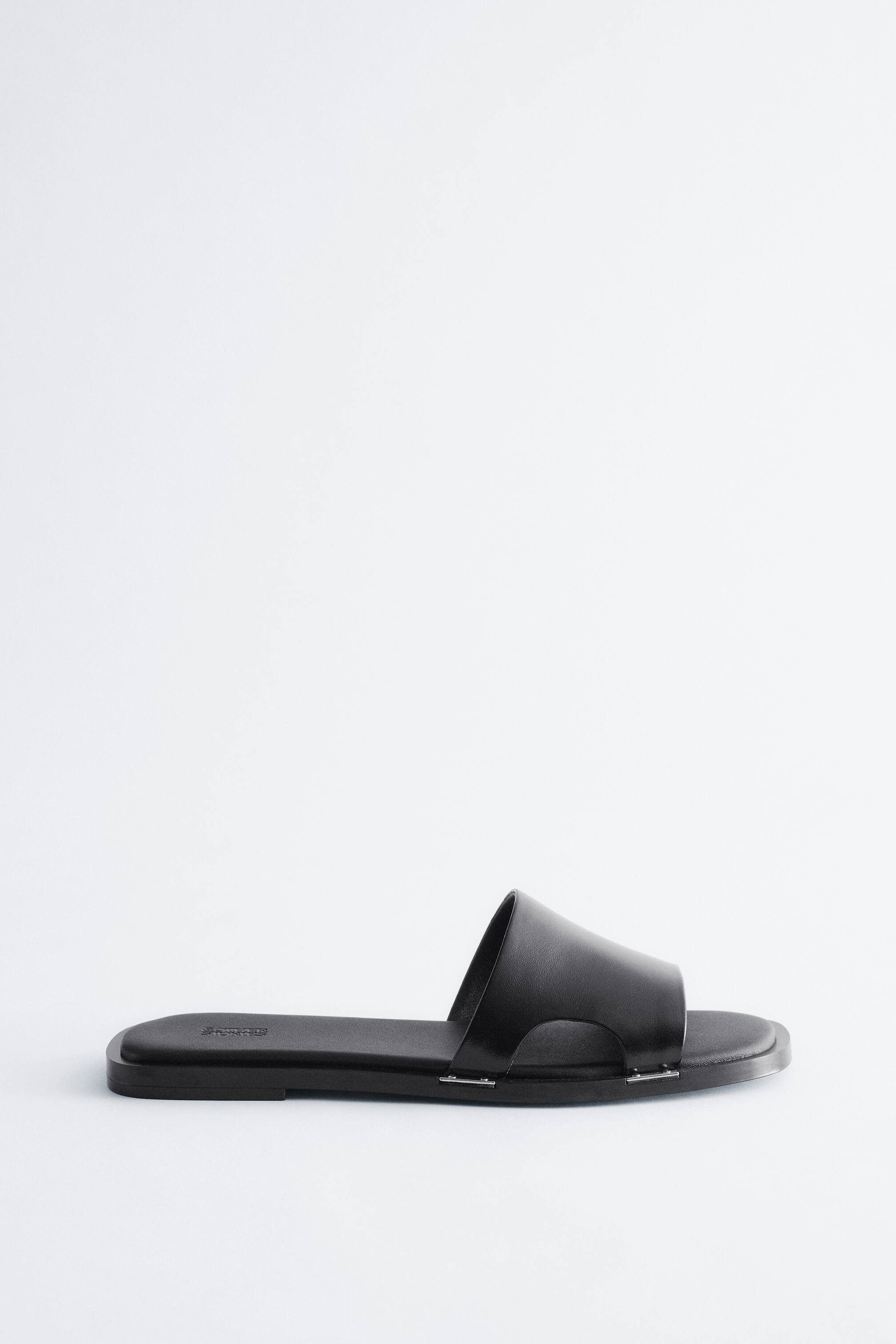 Προβολή μεγαλύτερης εικόνας: Ένα μαύρο σανδάλι με φαρδιά λωρίδα παρουσιάζει την αριστερή του πλευρά. Μια φαρδιά, λεία λωρίδα καμπυλώνεται πάνω από μια επίπεδη σόλα, ενώ δύο μικρές, ασημί μεταλλικές λεπτομέρειες συνδέουν τη λωρίδα με τη σόλα.