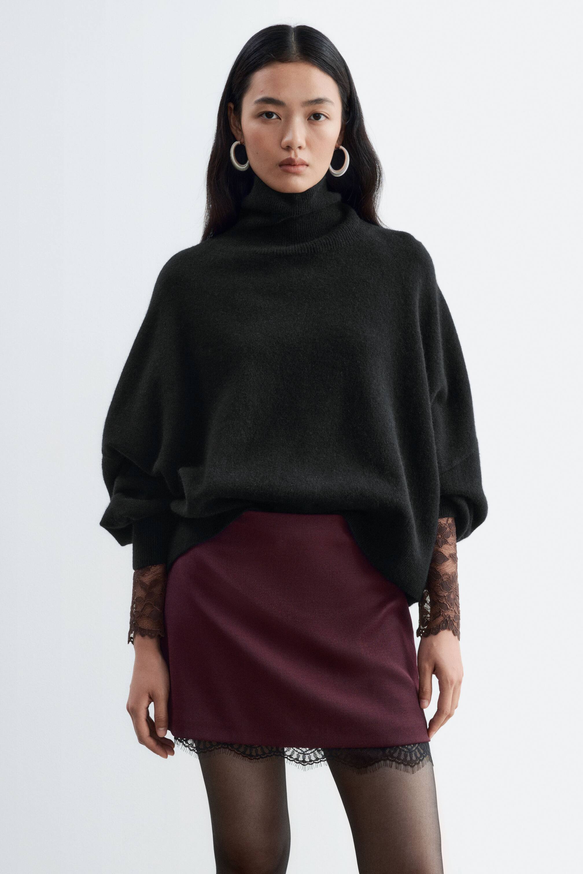 View larger image: A burgundy mini skirt featuring delicate black lace trim along the hem is being worn by the model. The skirt is styled with an oversized black roll-neck sweater and sheer black tights.