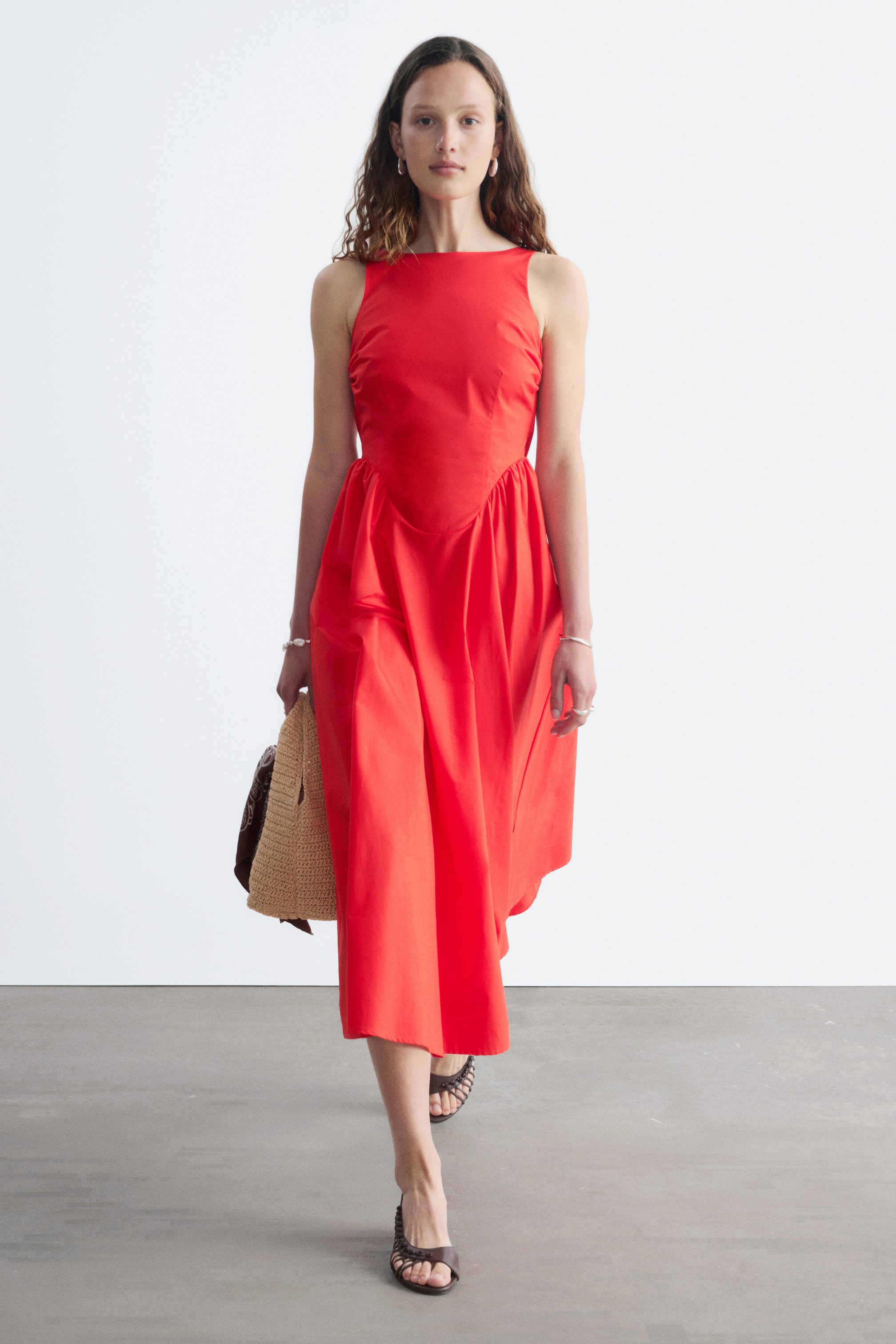 Vezi imaginea mai mare: O rochie midi roșie fără mâneci, cu un corsaj mulat cu pense frontale și o fustă evazată, plisată, este purtată de model. Modelul merge și poartă o geantă din paie împletite.