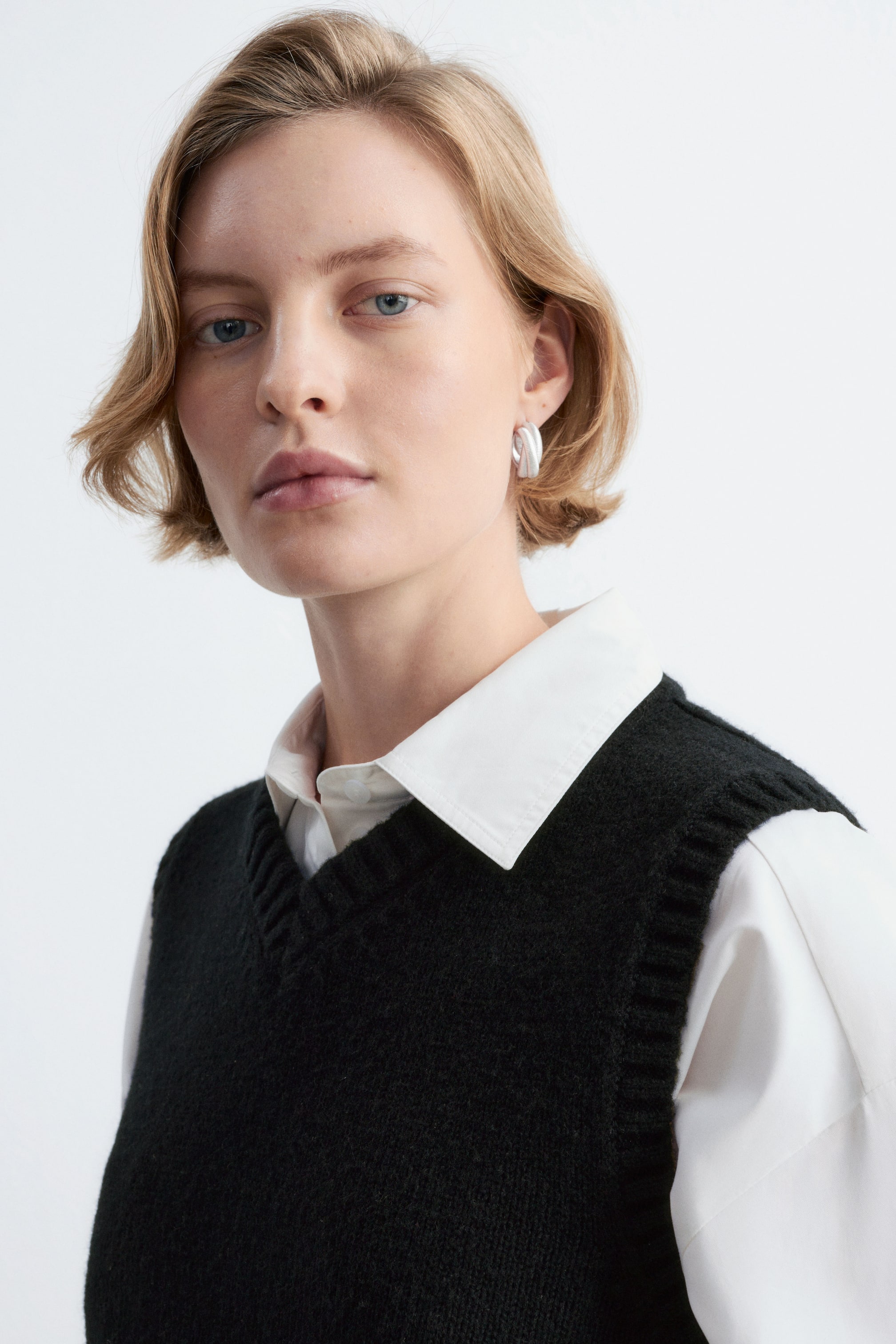 View larger image: A black wool-cotton knitted vest featuring a ribbed V-neck is worn by the model over a white collared shirt. A silver-toned plated brass hoop earring with a twisted, textured surface is also worn, visible on the model’s ear.