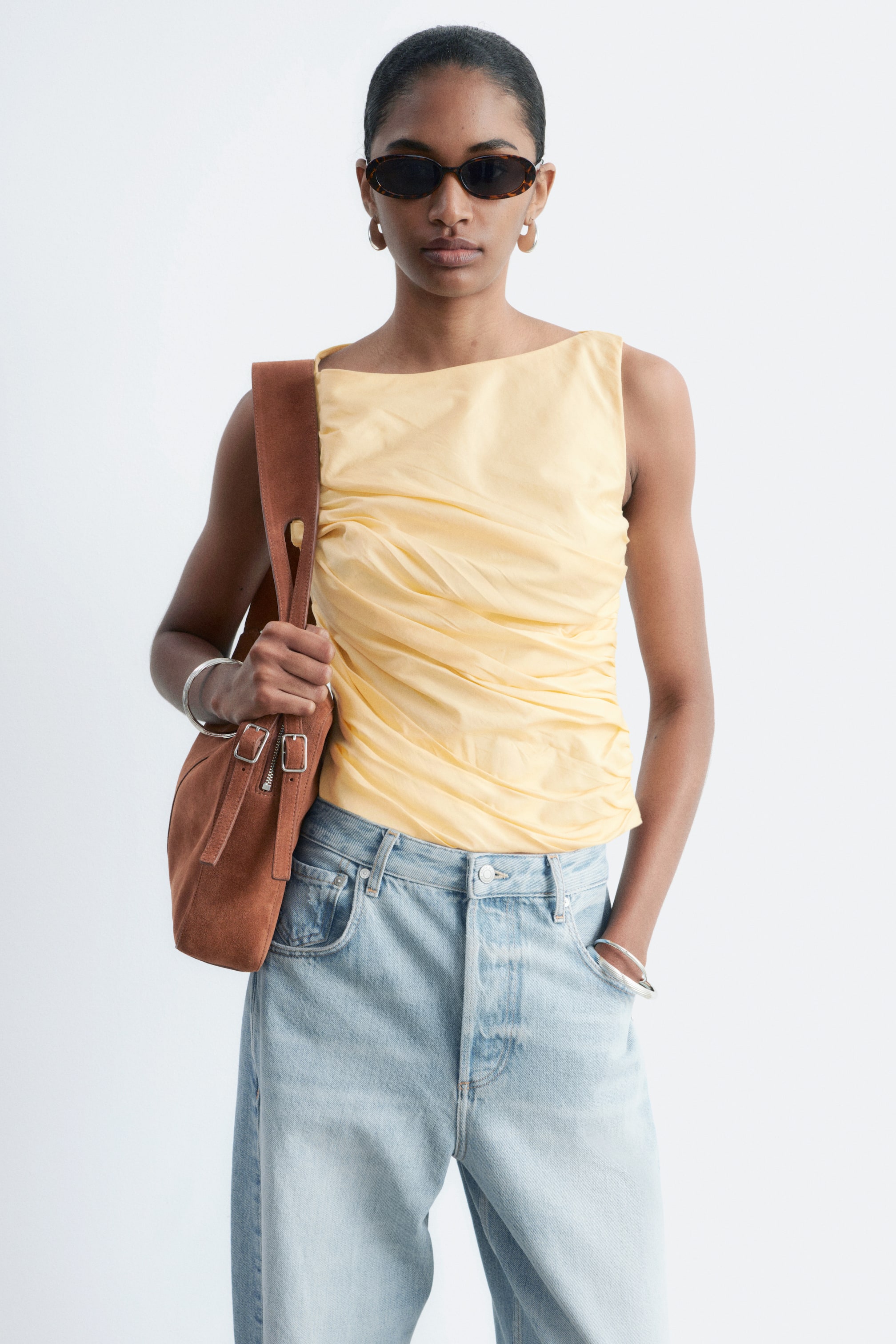 Visualizza immagine più grande: Un top senza maniche di colore giallo pallido presenta un ampio scollo e una vasta arricciatura che crea una texture drappeggiata sul davanti. Viene presentato con jeans a vita alta e lavaggio chiaro, che mostrano una vestibilità rilassata e una chiusura a bottoni visibile.