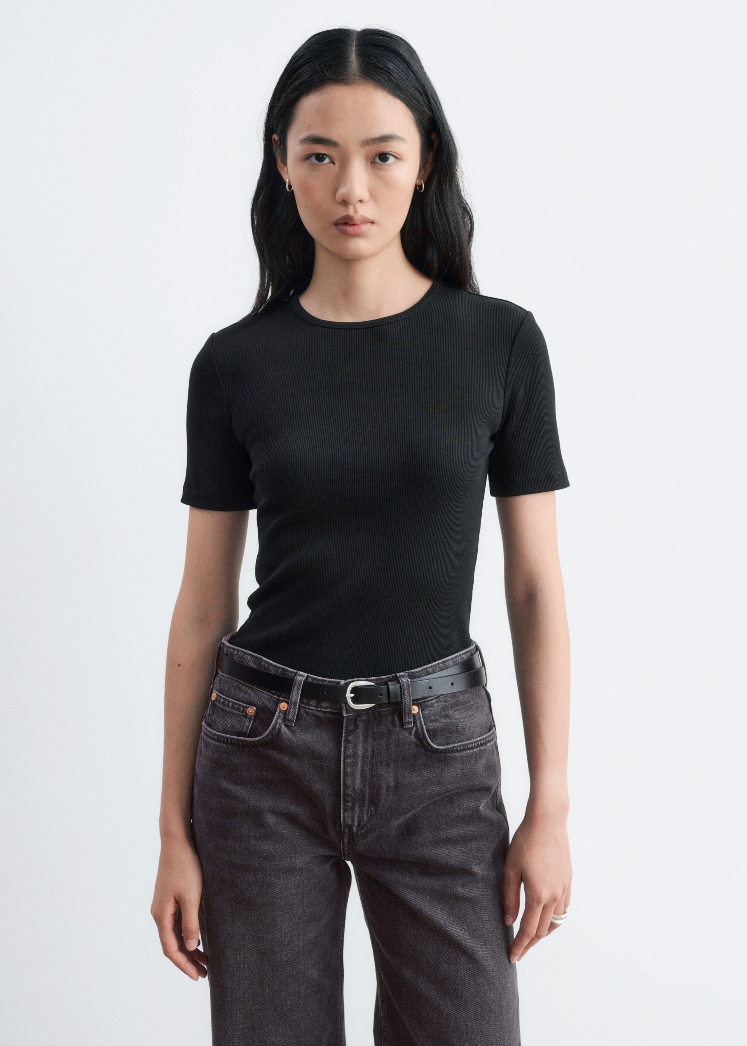 A black ribbed T-shirt featuring a round neck and short sleeves is being worn by the model, who is pairing it tucked into dark grey jeans with a black belt.
