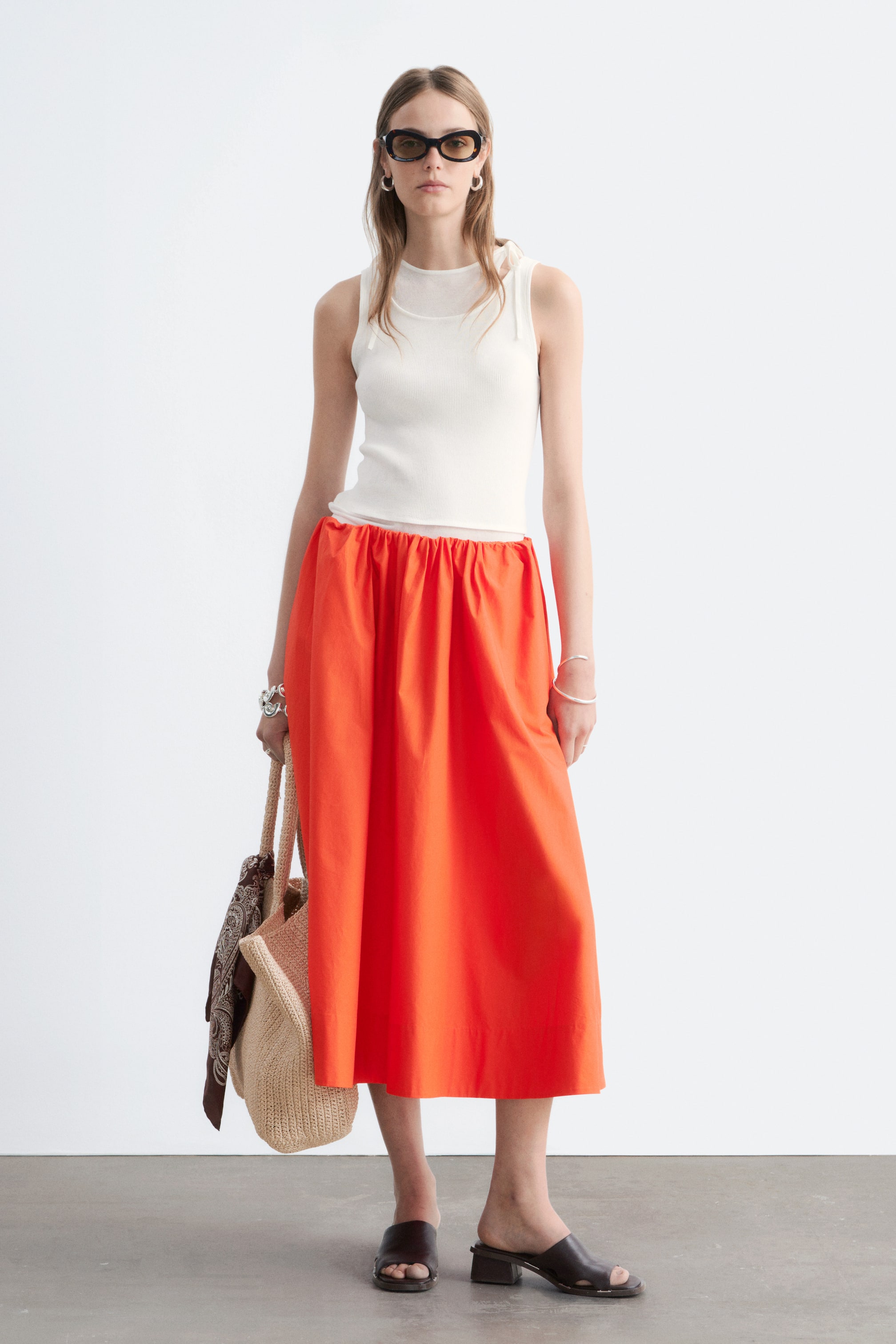 Agrandir l'image: La mannequin porte une jupe midi en coton orange, présentant une silhouette midi décontractée et une taille élastiquée reposant bas sur ses hanches. La mannequin associe la jupe avec un débardeur côtelé blanc, des mules foncées, et porte un sac fourre-tout en osier.
