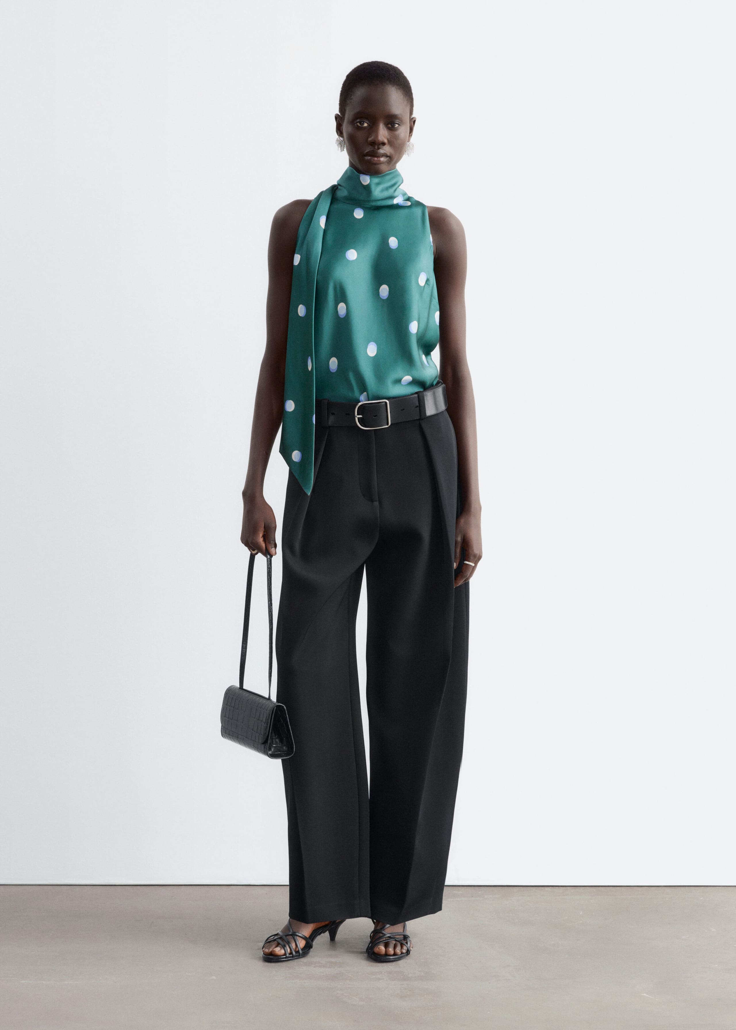 A green satin sleeveless top featuring a high neckline, a draped scarf detail, and a polka-dot pattern is worn by the model. The top is tucked into wide-leg black trousers styled with a black belt.