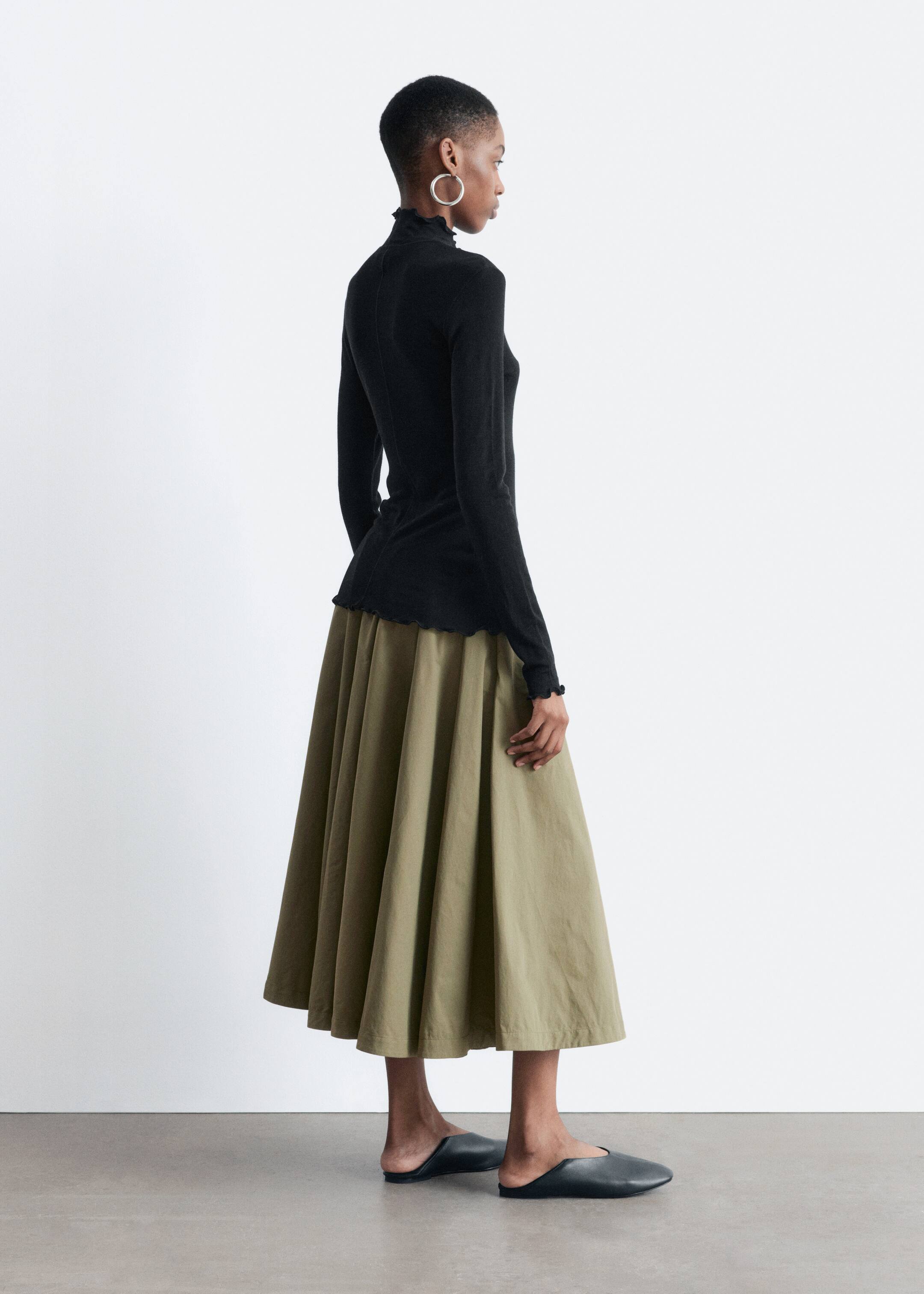 Vezi imaginea mai mare: Topul negru cu mânecă lungă, având un guler fals, margini ondulate și o cusătură plană în josul spatelui, este purtat de model. Bluza este stilizată cu o fustă plină verde măsliniu.