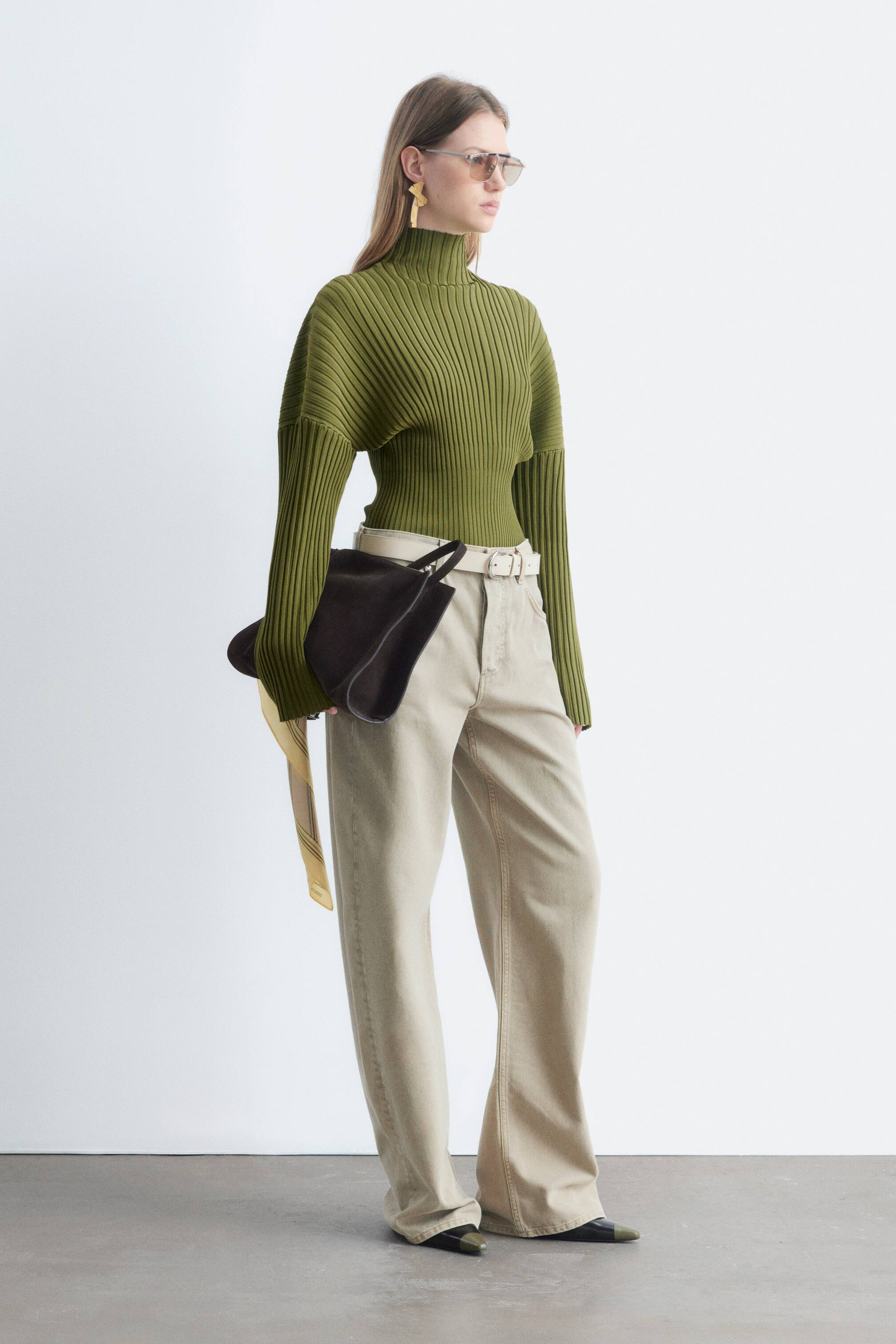 Agrandir l'image: Un pull à col montant simulé vert olive, doté de côtes bien marquées sur l’ensemble du vêtement, est porté par le mannequin. Le pull est assorti à un pantalon beige taille haute, et le mannequin porte un sac marron foncé.