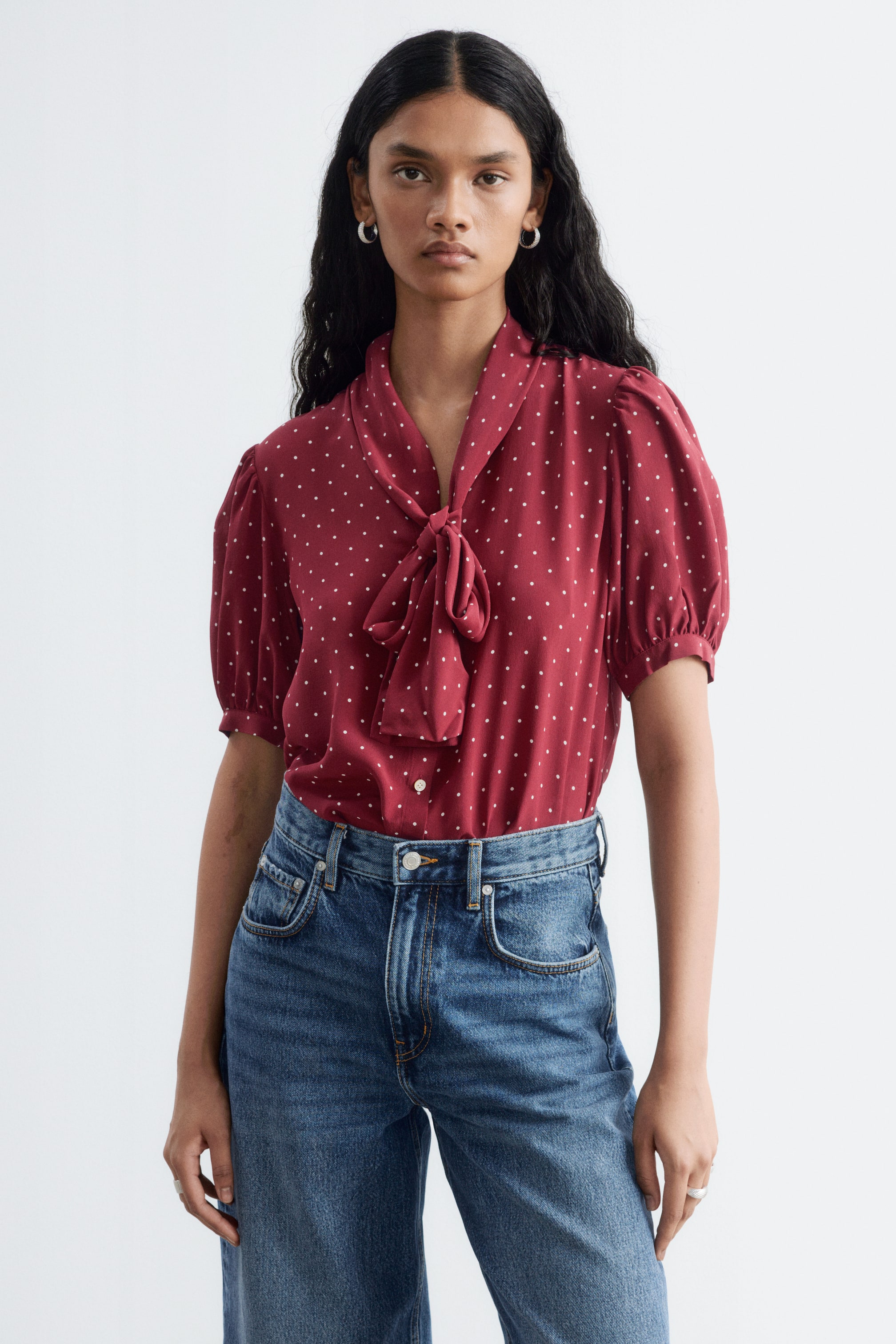 Ver imagen más grande: La modelo lleva una blusa de seda de color rojo oscuro con un estampado de microtopos blancos, metida por dentro de unos vaqueros azules de cintura alta. La blusa presenta mangas cortas abullonadas y un cuello anudado en forma de lazo.