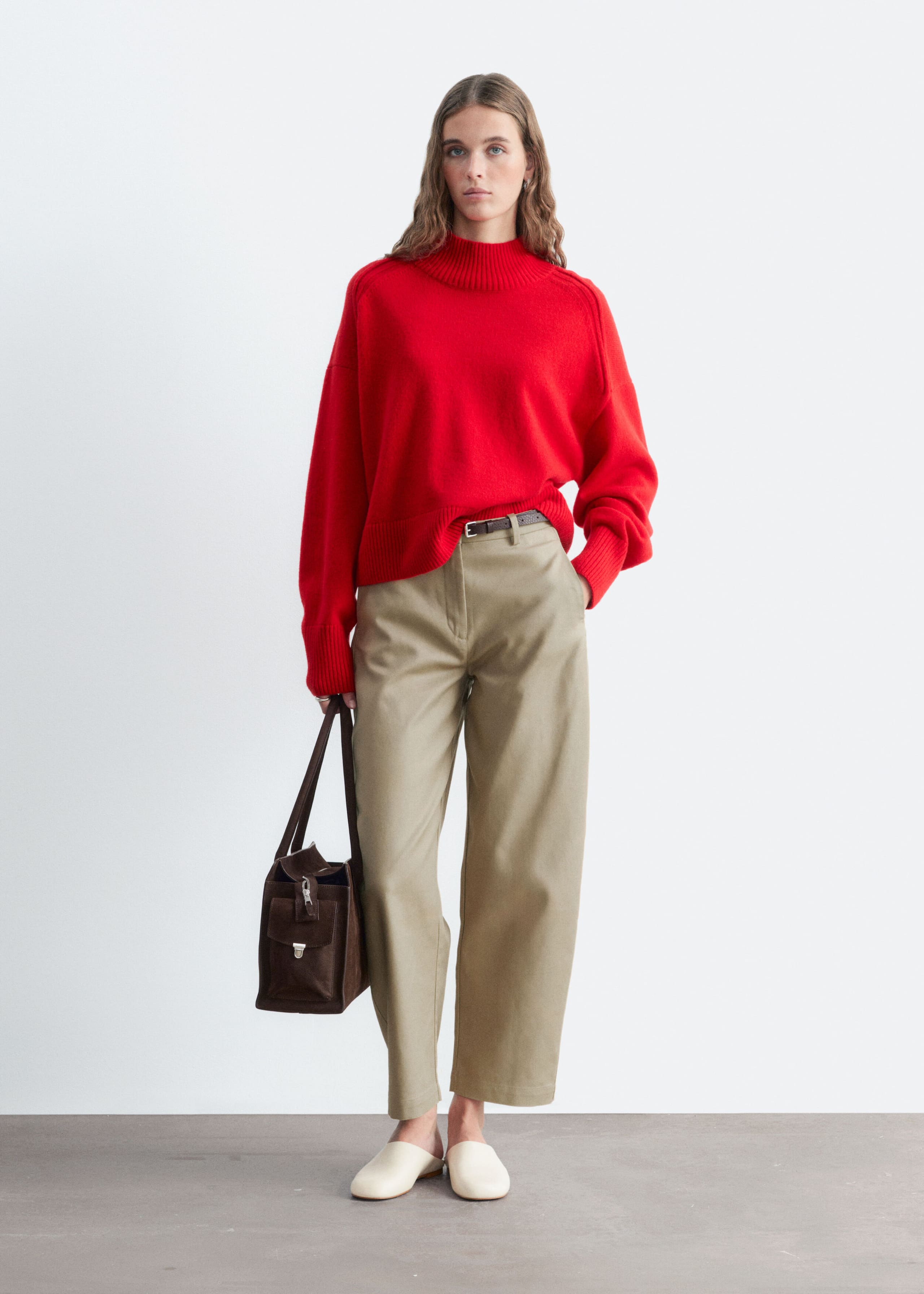 A red mock neck sweater featuring a ribbed collar, cuffs, and hem is worn by the model. The sweater has a relaxed silhouette and is styled with a partial tuck into a pair of beige trousers.