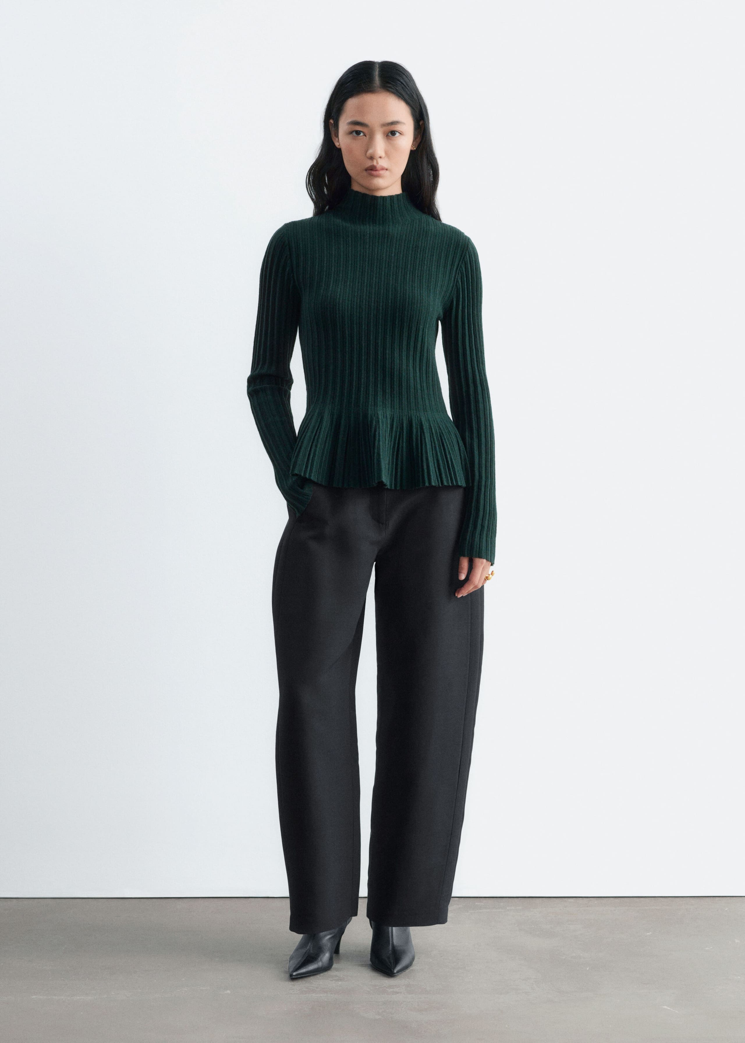 The model is wearing a dark green ribbed-knit top featuring a mock neck, long sleeves, and a flared peplum hem. The product is styled with black wide-leg trousers and black pointed-toe boots.
