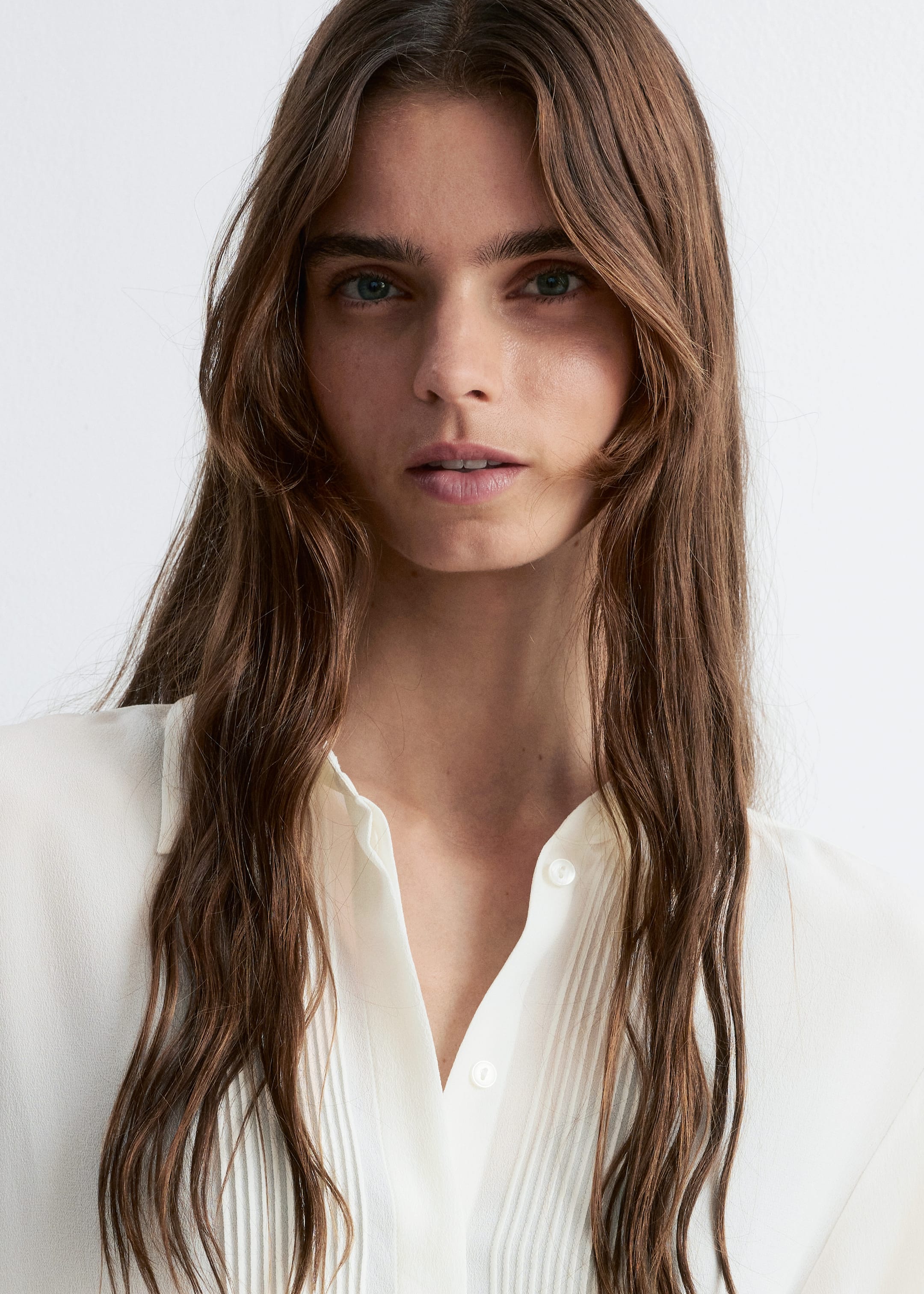 Grotere afbeelding bekijken: Een off-white blouse wordt gepresenteerd vanuit een vooraanzicht. Het heeft een puntige kraag, met de bovenste knoop los, en een centrale knopenlijst. Verticale plooien sieren de voorpanden van het kledingstuk.