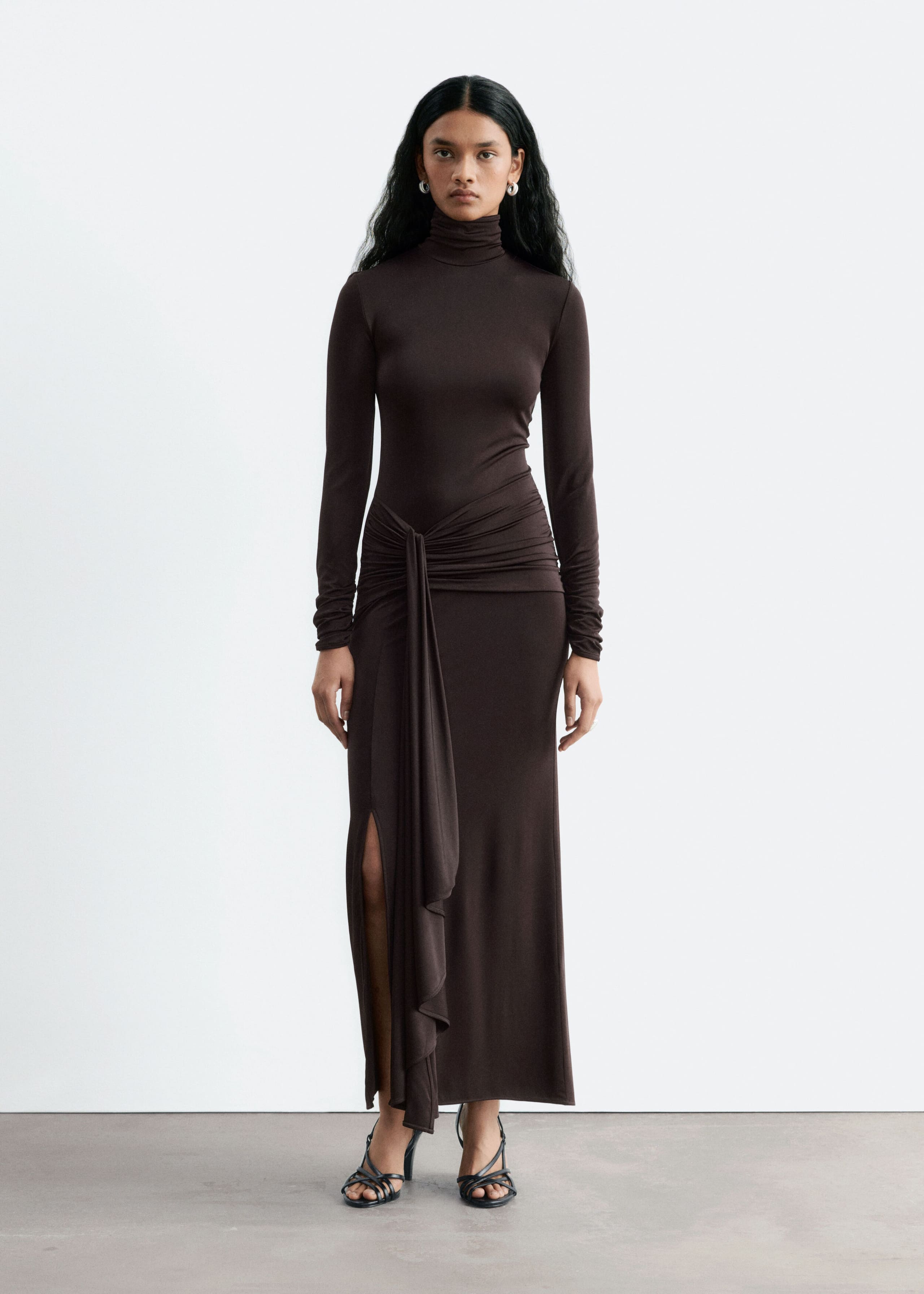 A dark brown jersey maxi dress is worn by the model, featuring a high mock neck, long sleeves and a slim-fit silhouette. The dress is designed with a gathered, draped tie detail at the waist that falls down the front next to a side leg split, and is styled with black strappy high-heeled sandals.