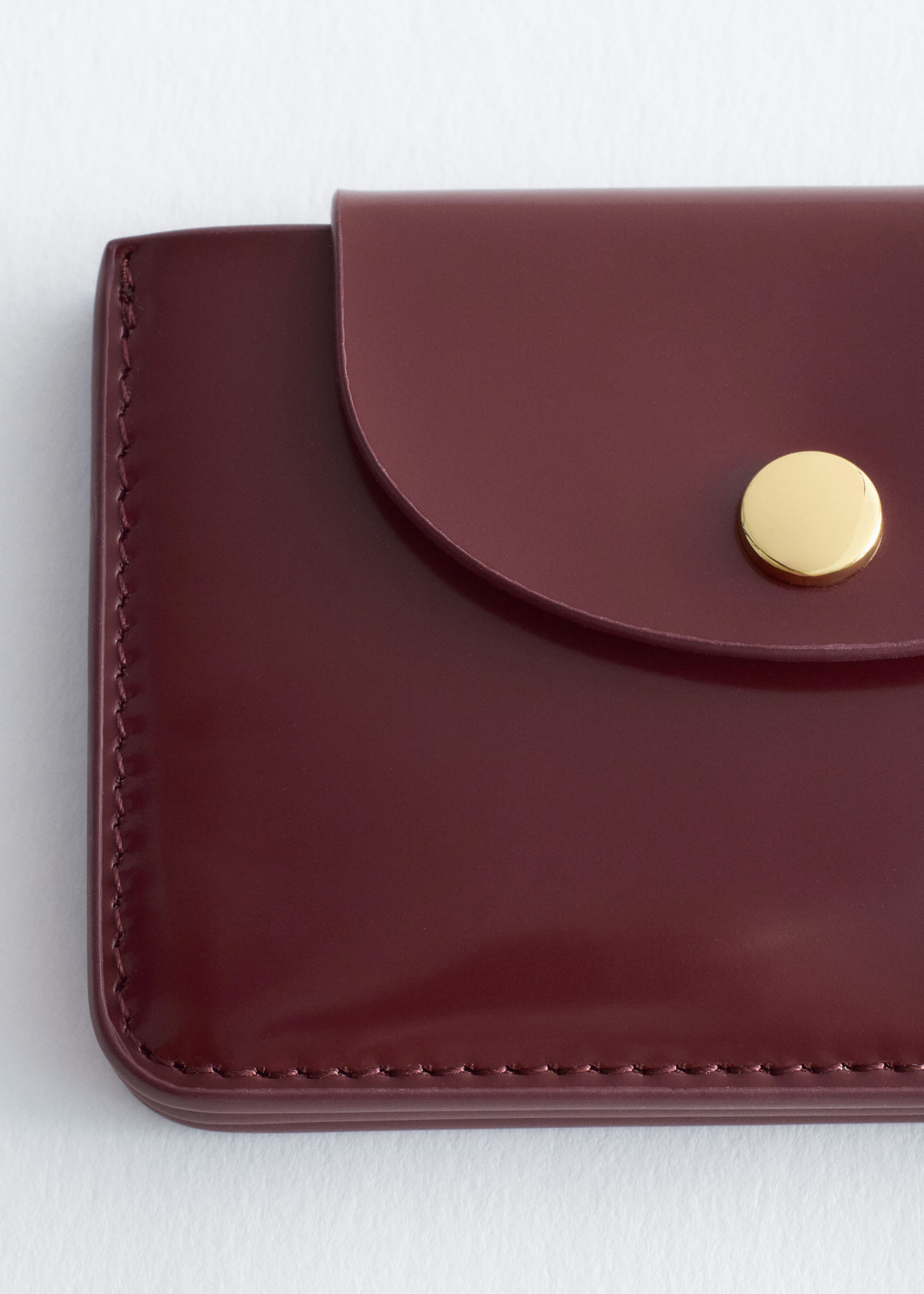 A burgundy leather cardholder is shown in a close-up front view, featuring a curved flap with a gold-tone circular snap closure. The cardholder features tonal stitching along its smooth, structured edges.