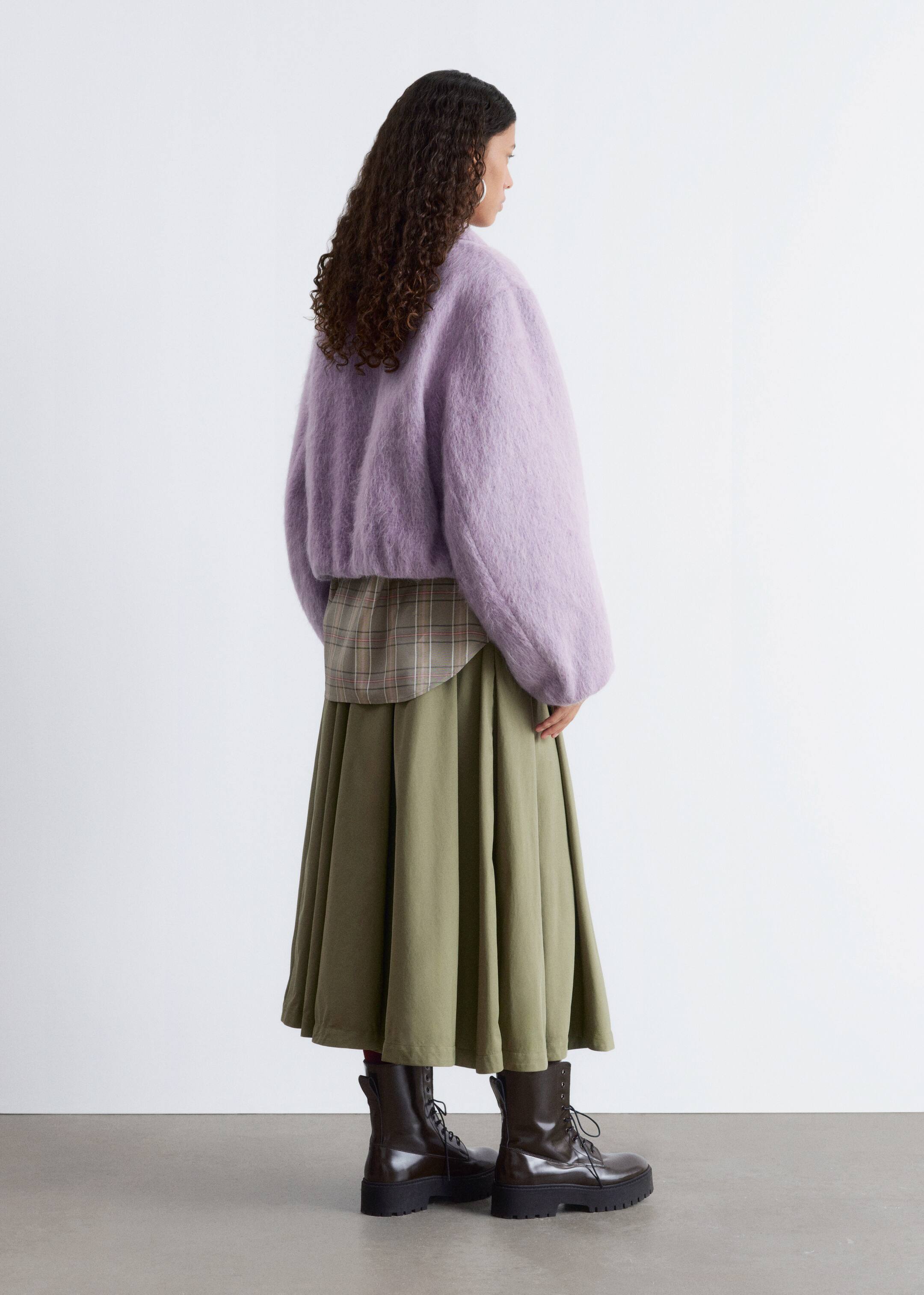 Visualizza immagine più grande: Un maglione tagliato a mo' di peluche color lilla è stratificato sopra una camicia a quadri, con il suo orlo curvo che si mostra sotto. Una lunga gonna a pieghe verde oliva cade fino alle caviglie, abbinata a stivali da combattimento stringati marrone scuro, il tutto visto da dietro a destra.