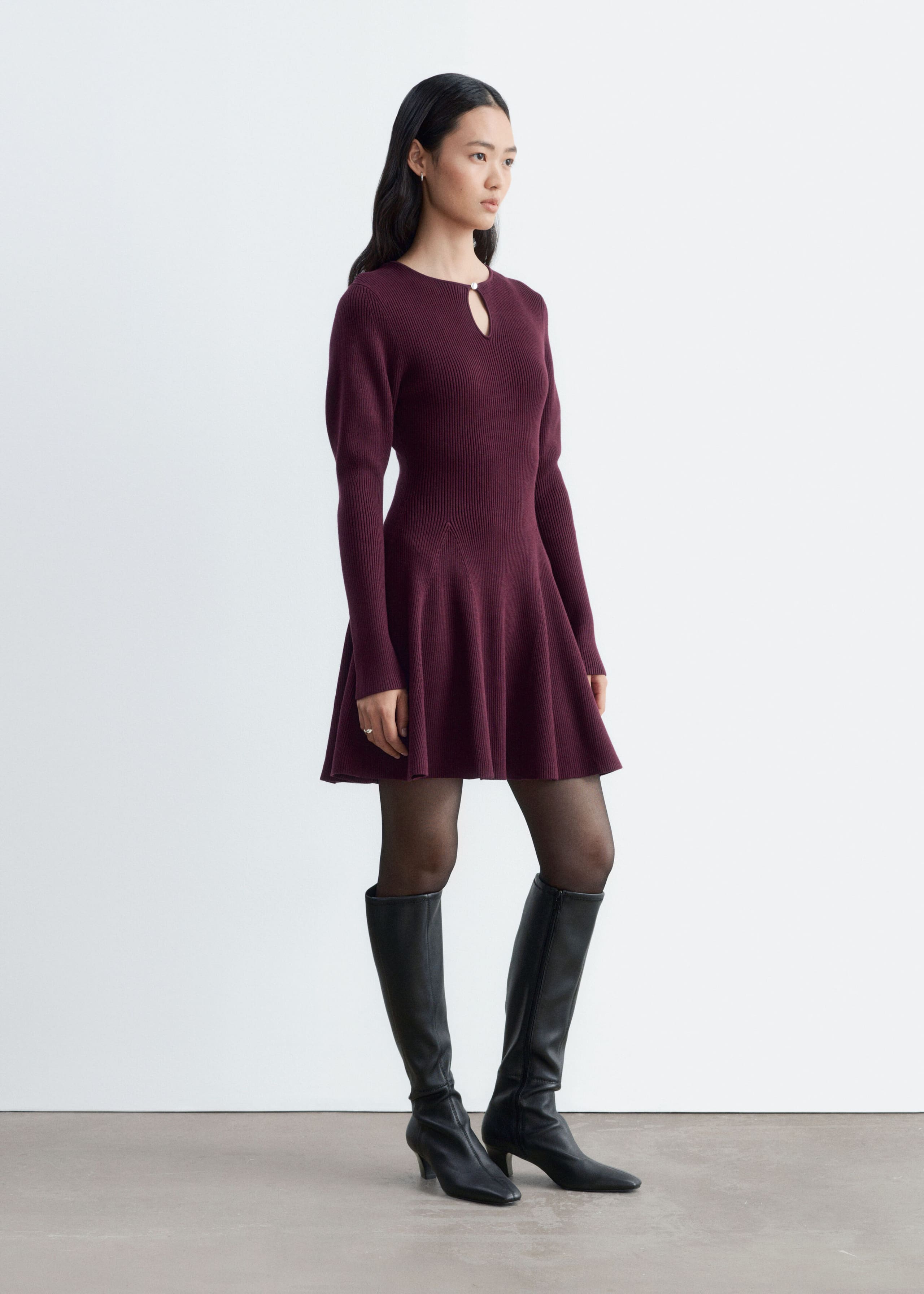 A ribbed knit long-sleeve dress in a deep burgundy hue is worn by the model, featuring a keyhole neckline with a metal button and a flared paneled skirt. The dress is styled with sheer black tights and black leather knee-high boots.