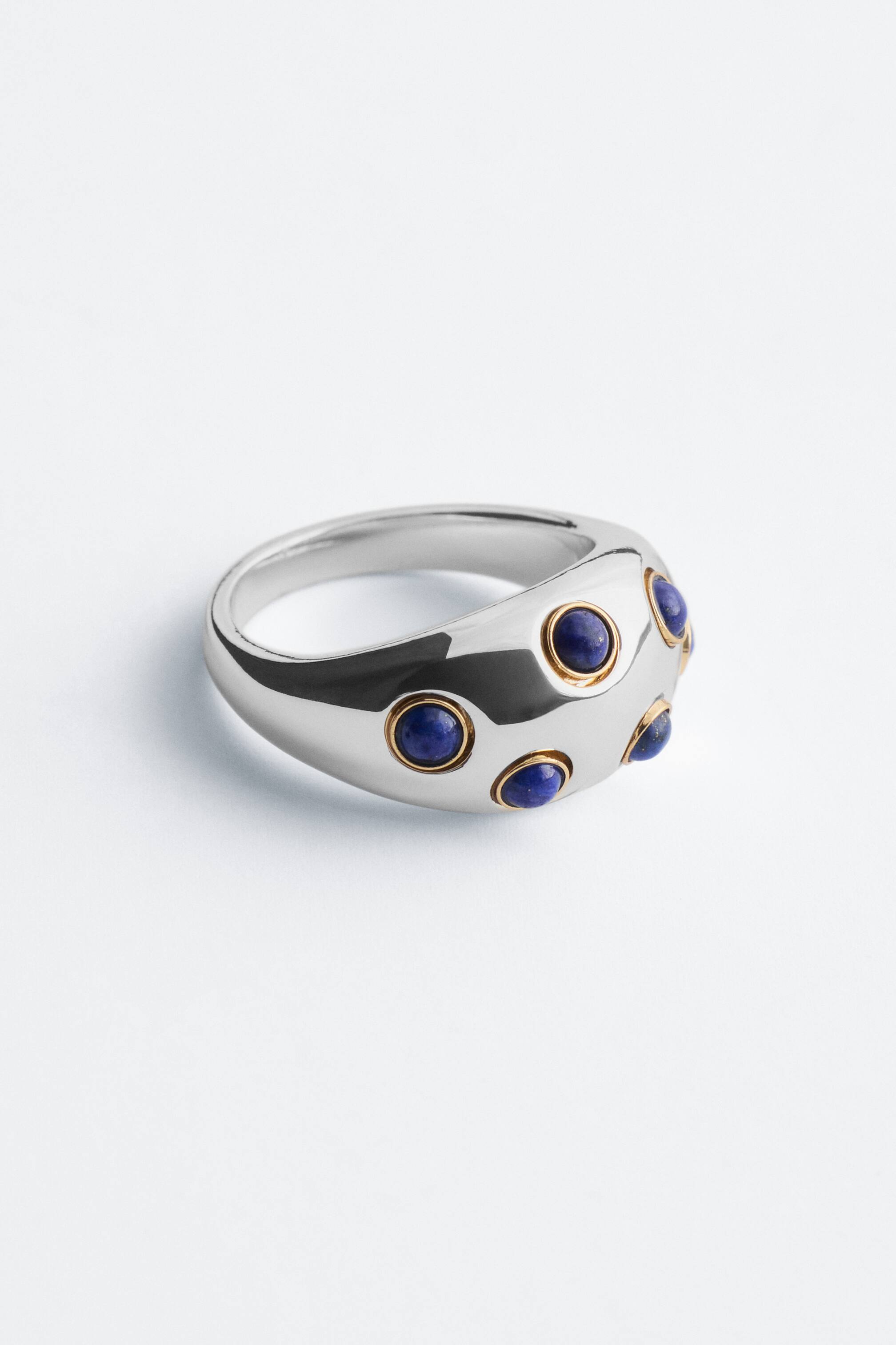 Grösseres Bild ansehen: Ein polierter, silberfarbener, skulpturaler Dome-Ring liegt in einer Dreiviertelansicht und zeigt mehrere runde blaue Cabochon-Steine, die in goldfarbenen Fassungen auf der gewölbten Oberfläche verteilt sind.