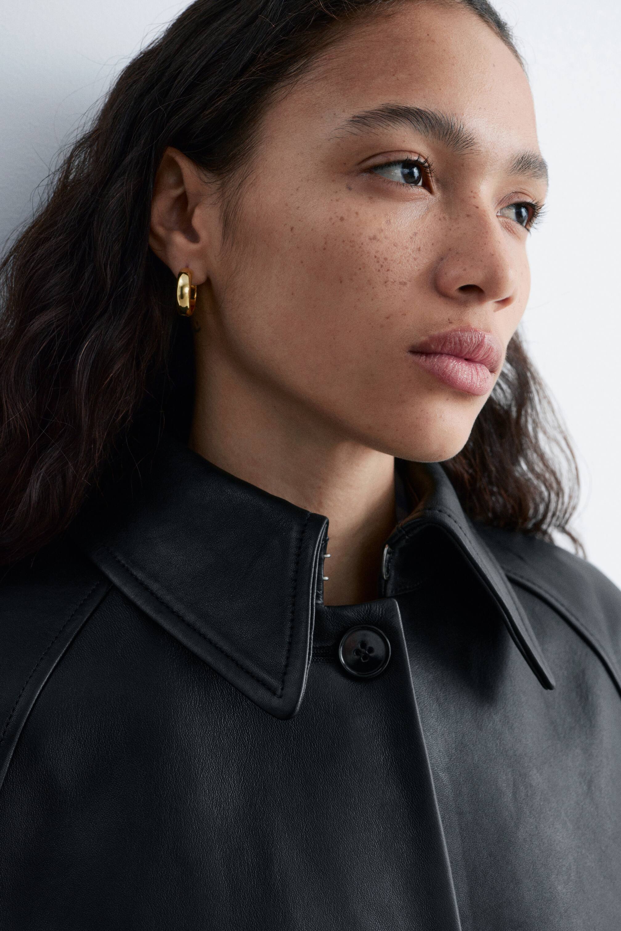 Vis større bilde: En svart skinnjakke med en strukturert spiss krage, en enkelt knappe-lukking foran, og synlige sømdetaljer på skuldrene. Et tykt, glatt gulløredobb pryder øreflippen.