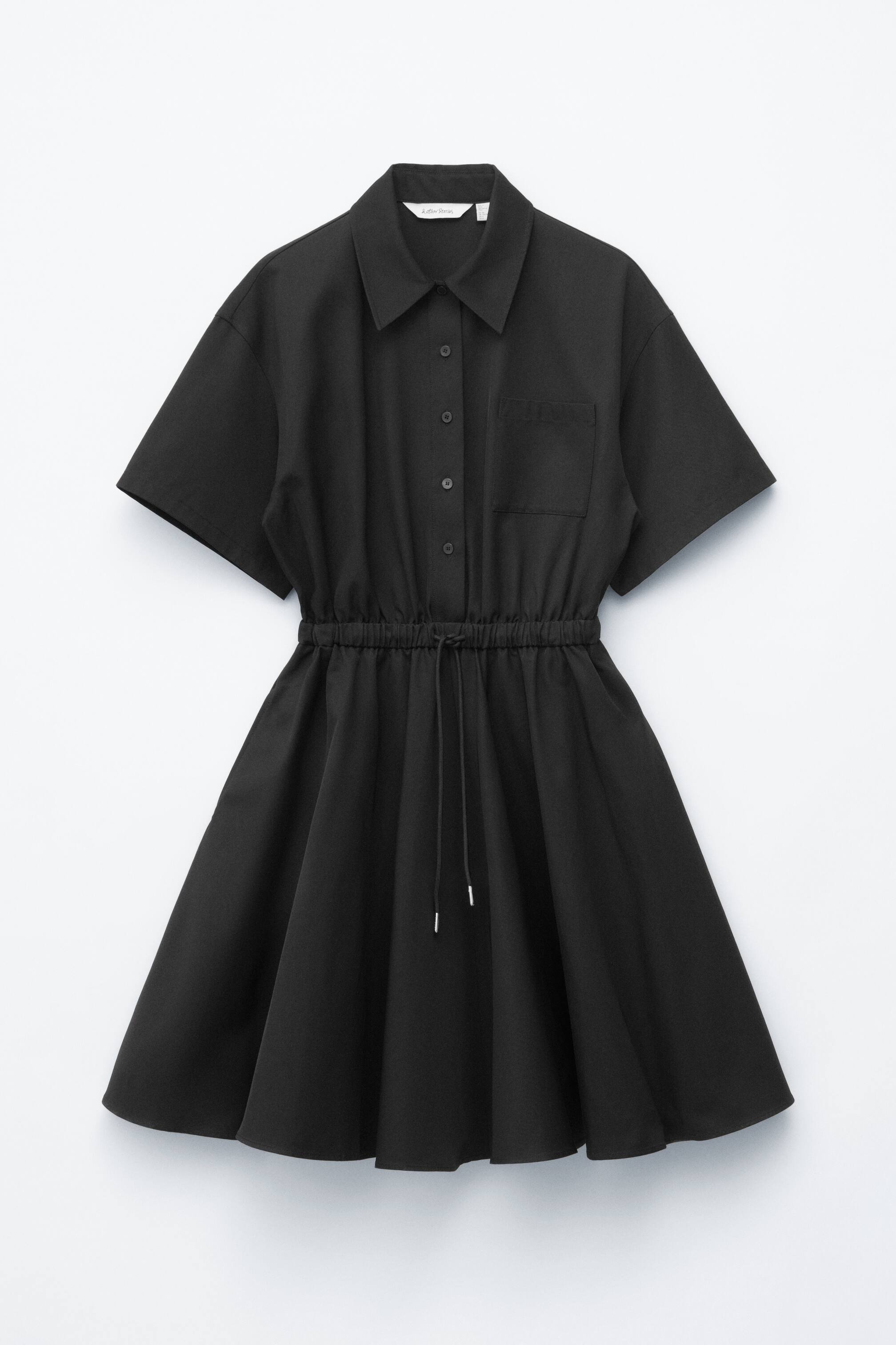 Agrandir l'image: Une robe chemise noire est présentée de FACE, avec un col classique, des manches courtes, des boutons ton sur ton sur le devant, une poche poitrine unique, une taille froncée avec cordon de serrage noir et embouts argentés, et une jupe évasée.