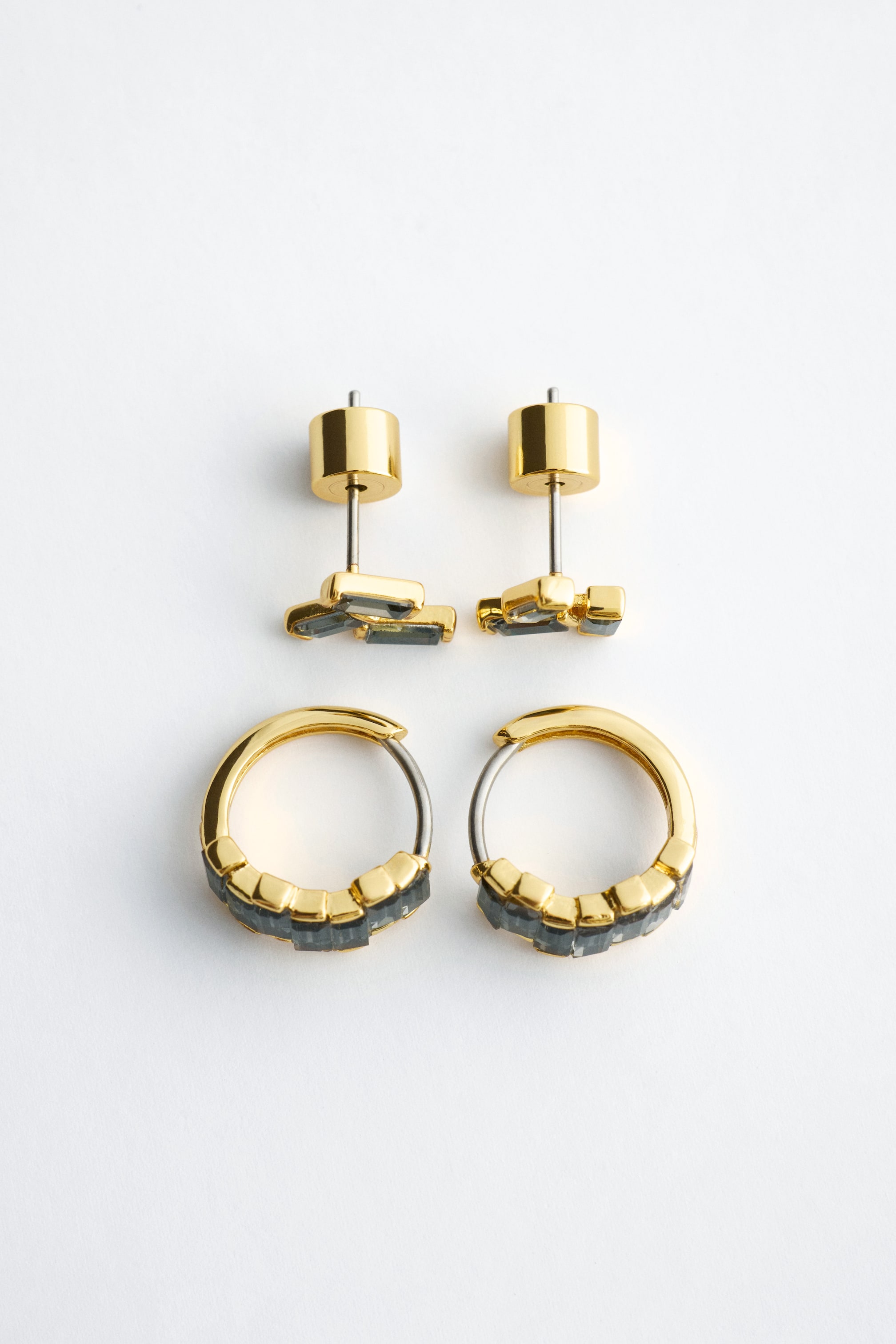 View larger image: A gold-toned brass earring set is lying flat, featuring a pair of small hoop earrings and a pair of clustered stud earrings facing forwards. Both pairs feature dark grey, rectangular-cut cubic zirconia stones set within the gold-toned frames.
