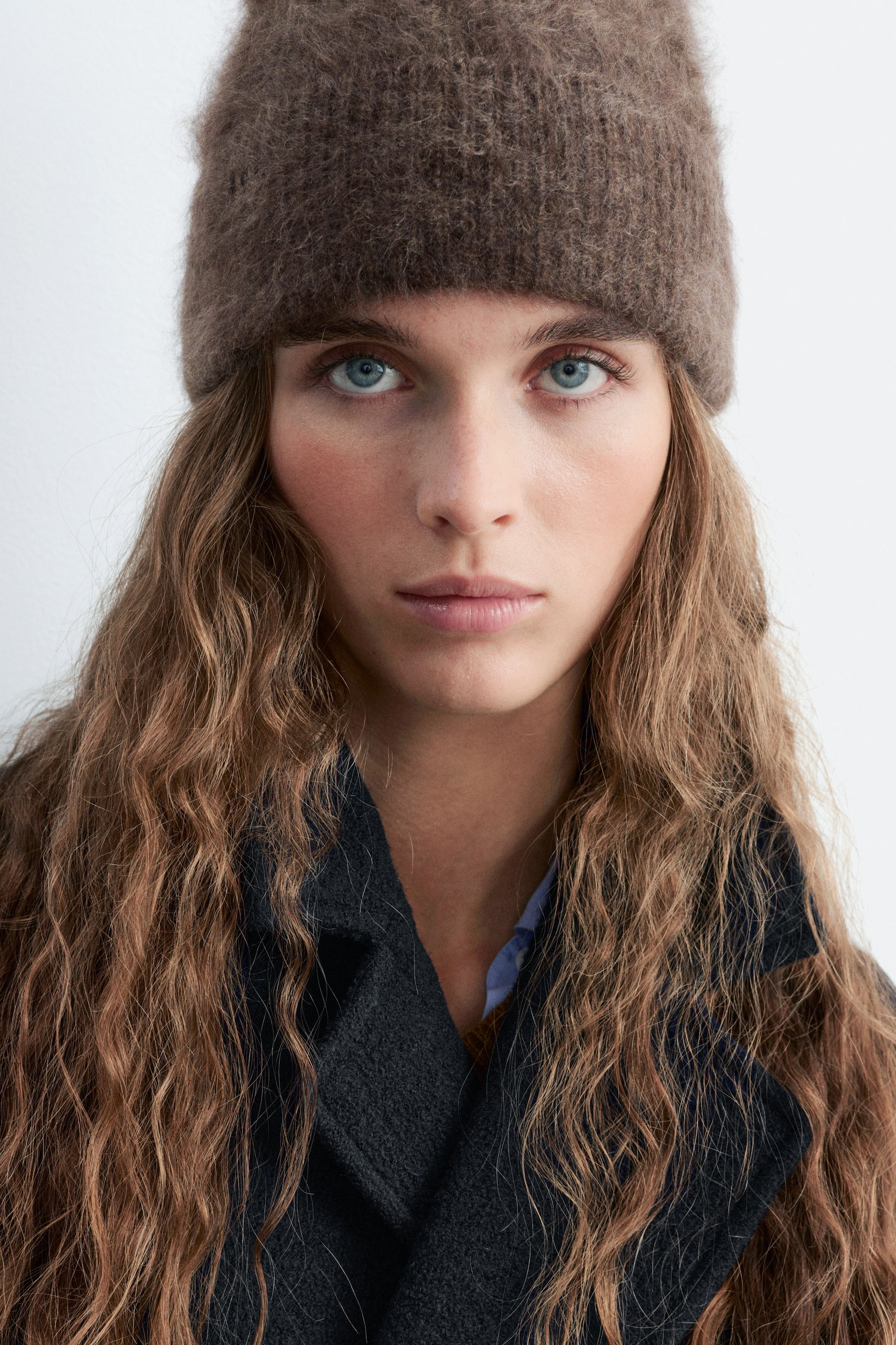 Agrandir l'image: Un bonnet en maille brun duveteux est porté par le mannequin, posé bas sur le front. Le bonnet est assorti à un manteau en laine gris foncé et à une chemise bleue claire à col.