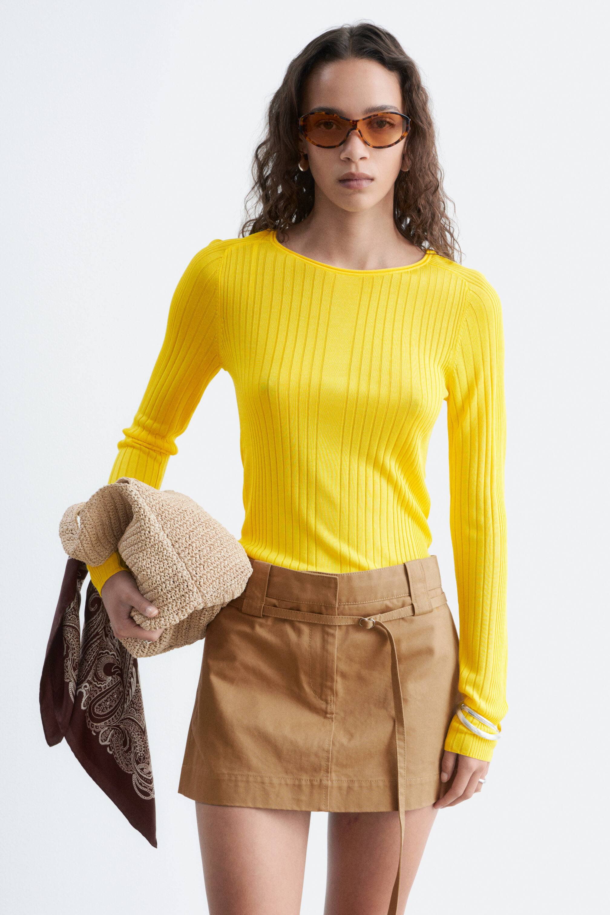 Grotere afbeelding bekijken: Een geel ribgebreid topje met lange mouwen wordt gedragen door het model. Het heeft een slank silhouet en een ronde halslijn met een omgeslagen rand. Het model draagt ook een beige minirok en houdt een geweven tas vast met een bruin patroon sjaal.