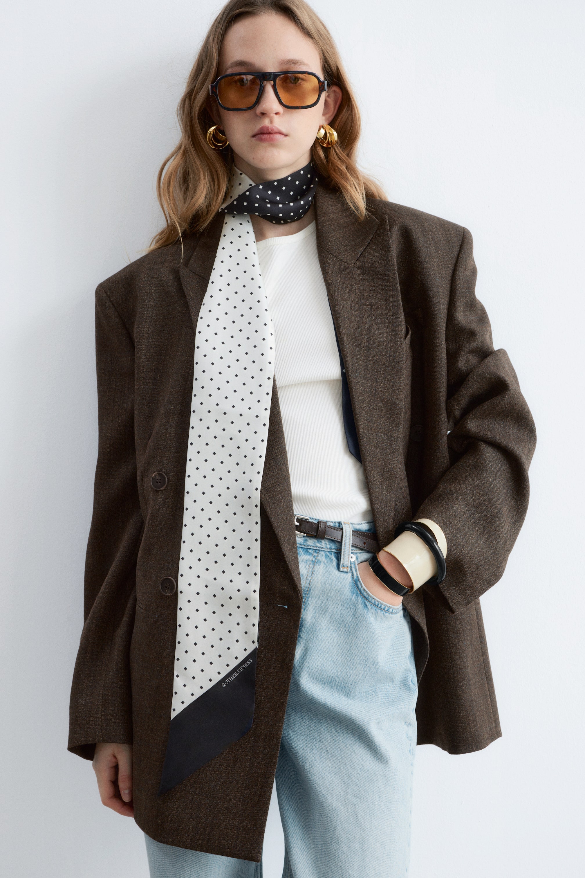 Agrandir l'image: Une longue écharpe est portée, avec un motif à pois noirs au niveau du cou et un motif à pois blancs sur la partie pendante, se terminant par une pointe noire. Cette écharpe est superposée à un blazer oversize brun foncé avec des revers crantés, qui recouvre un haut côtelé blanc et un jean délavé clair.