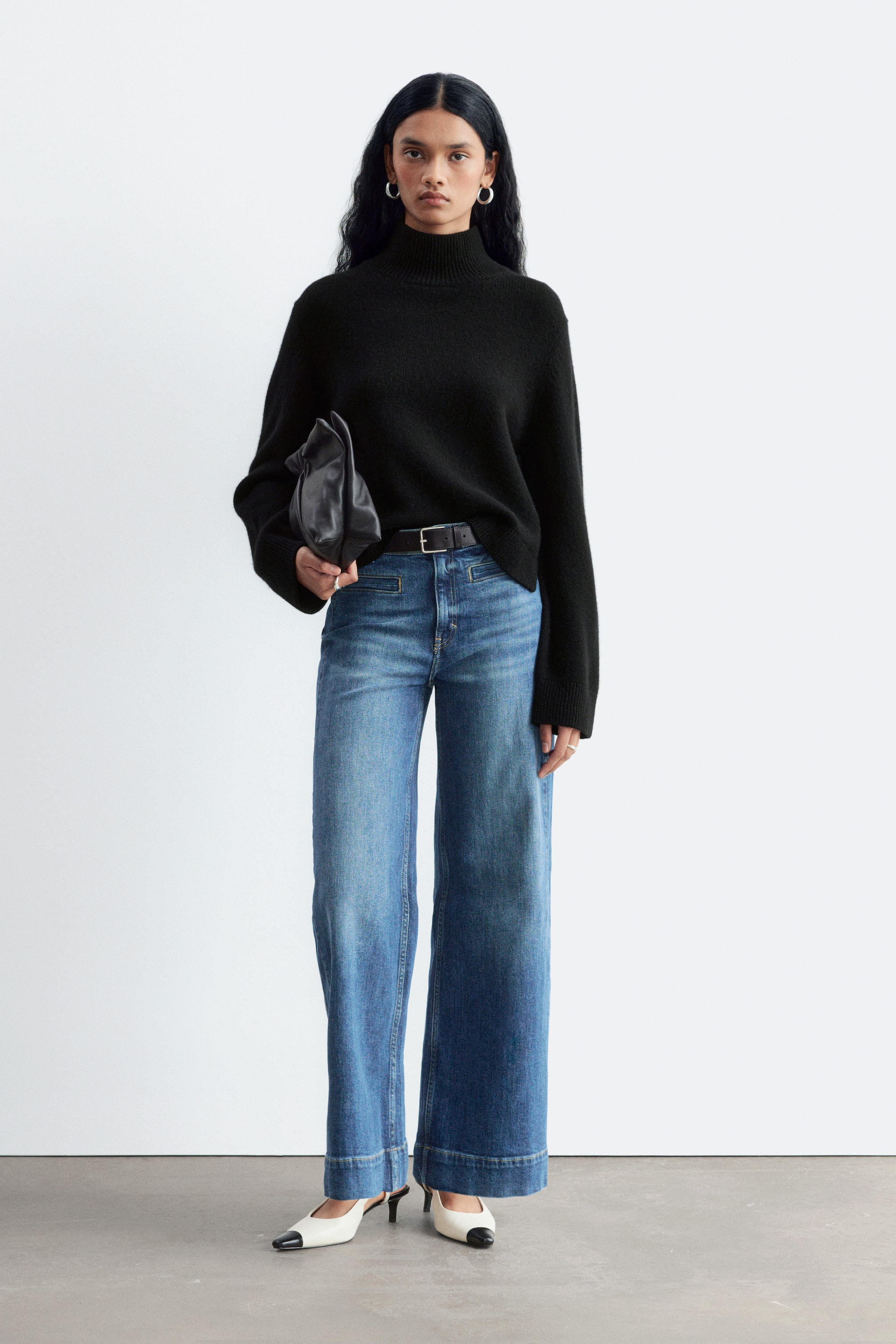 Agrandir l'image: Le mannequin porte un jean regular à jambes larges dans un délavage bleu moyen, avec une taille haute et un léger effet délavé le long des cuisses. Le jean est associé à une ceinture en cuir noire, un pull noir à col montant rentré à l’intérieur, et des escarpins bicolores à bout pointu.