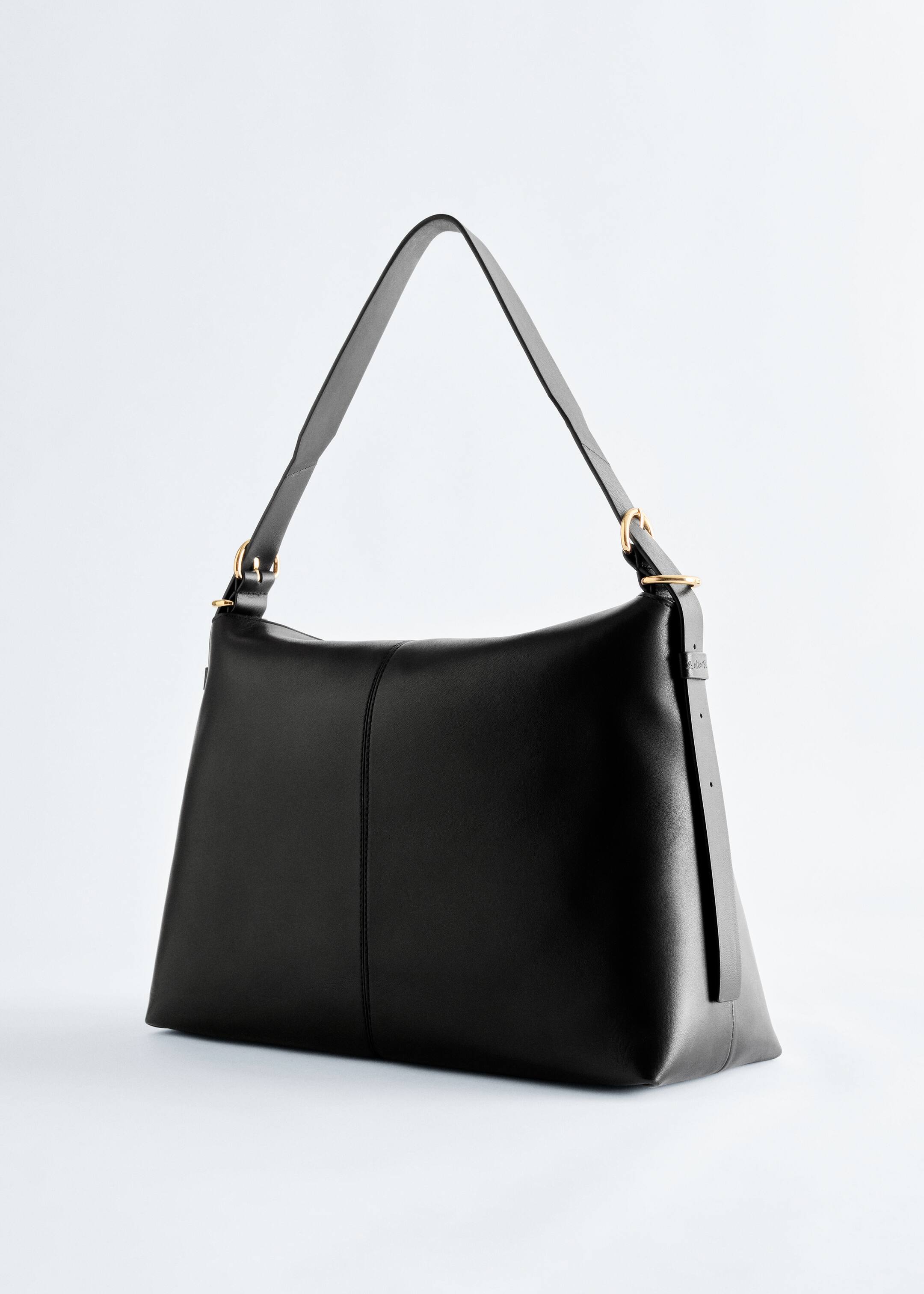 Grösseres Bild ansehen: Eine schwarze Lederschultertasche wird in einem bestimmten Winkel präsentiert und weist eine elegante Form mit einer vertikalen Naht auf, die in der Mitte ihrer Vorderseite verläuft. Ein breiter, verstellbarer Schulterriemen ist mit goldfarbenen Ringen und Schnallen an der Tasche befestigt.