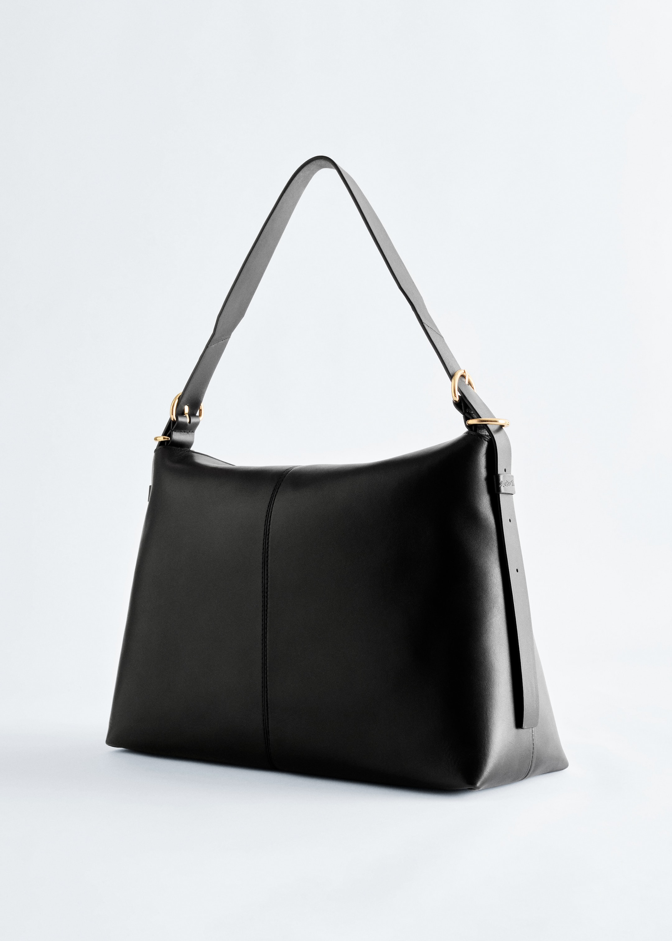 Größeres Bild ansehen: Eine schwarze Lederschultertasche wird in einem Winkel präsentiert und zeichnet sich durch eine schlanke Form mit einer vertikalen Naht an der Vorderseite aus. Ein breiter verstellbarer Schulterriemen ist mit goldfarbenen Ringen und Schnallen an der Tasche befestigt.